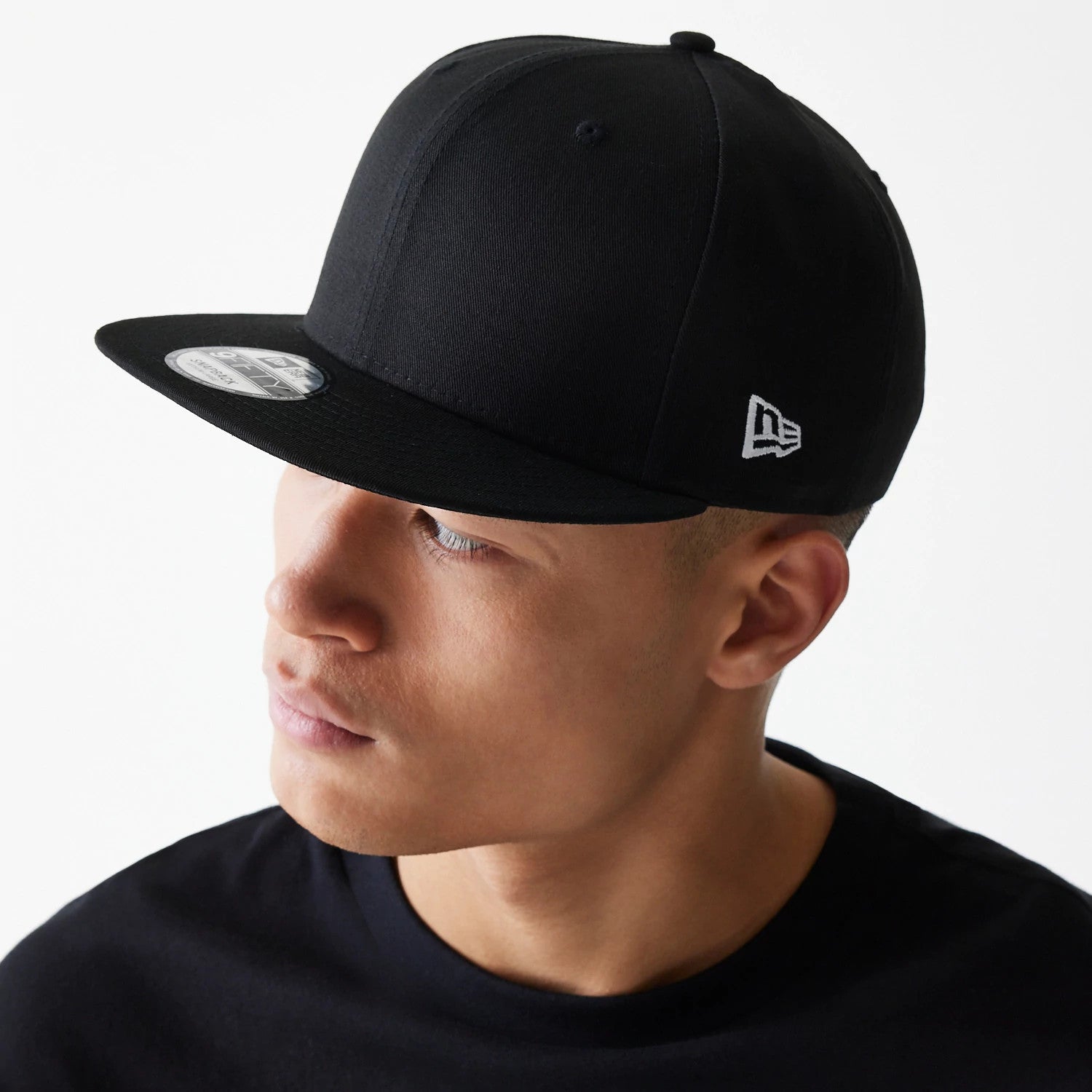 New Era All Black 9FIFTY Cap, three-quarter view on a model showing the grey embroidered New Era flag logo on the left side, flat black visor with a 9FIFTY size sticker, and structured crown.