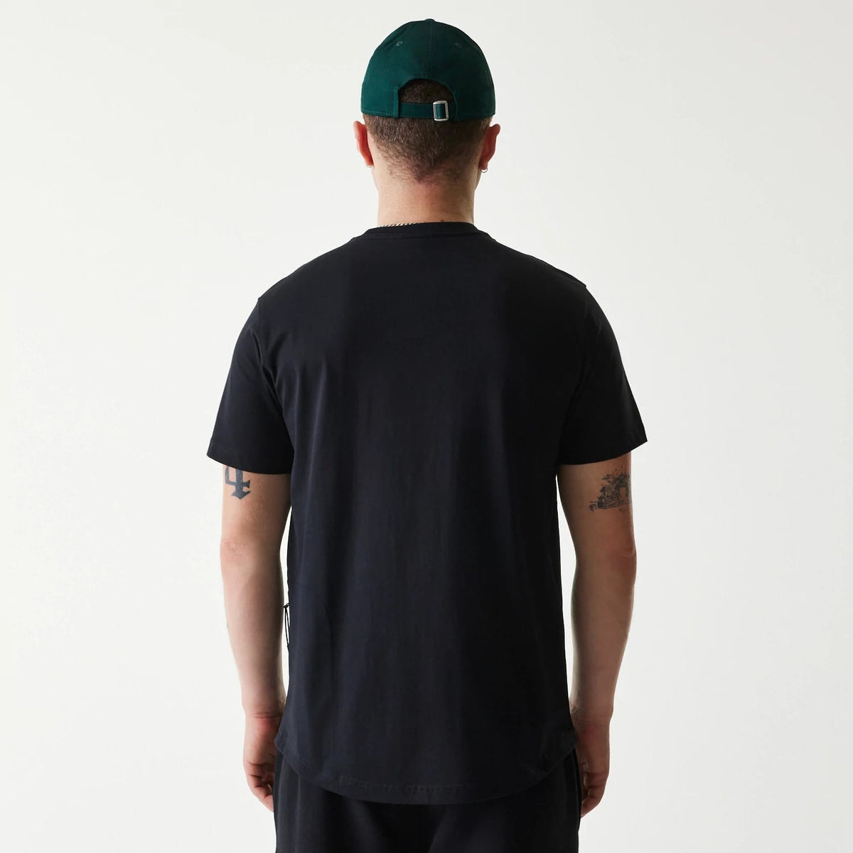 New Era Outdoor Earth Day Black T-Shirt, on-model rear view showing the plain black fabric extending over the shoulders, hem finishing below the waist, and a green cap with a debossed New Era logo on the back of the head.