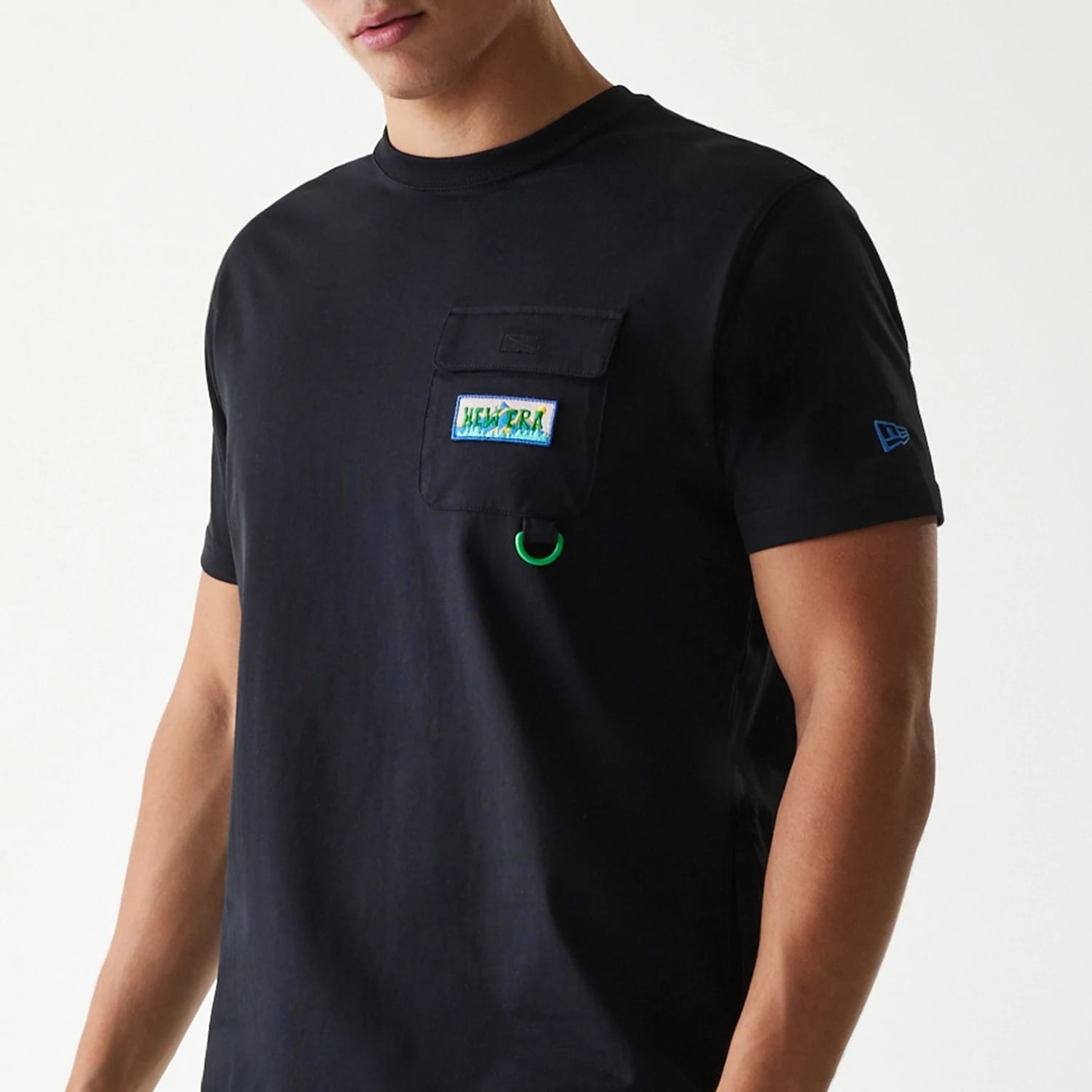 New Era Outdoor Earth Day Black Side Pocket T-Shirt, front view showing a black T-shirt with a small flap pocket on the chest featuring an embossed New Era logo, colourful patch, green ring below, and a blue New Era logo on the left sleeve.