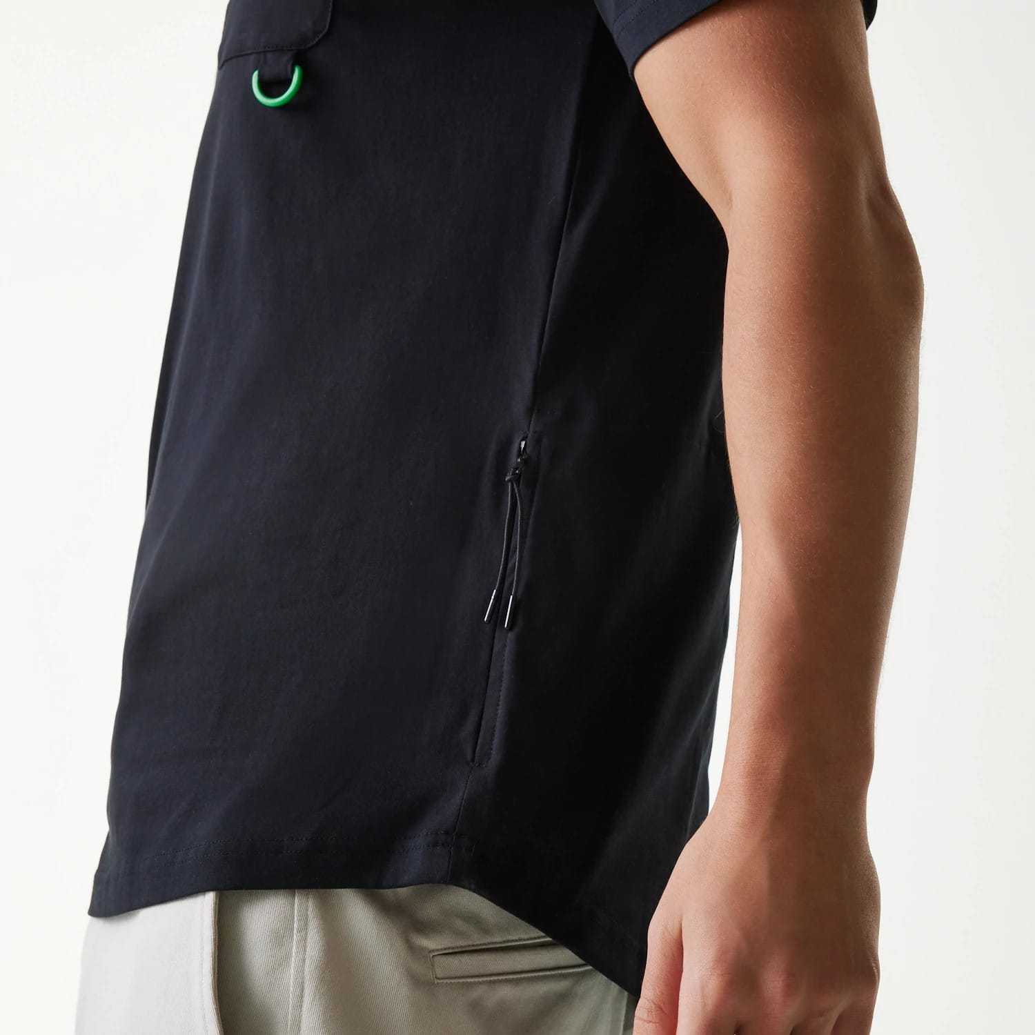New Era Outdoor Earth Day Black Side Pocket T-Shirt, side view showing a sleek black design with a zip pocket, short sleeves ending just above the elbow, a small green loop near the shoulder and the hem sitting above light-coloured trousers.