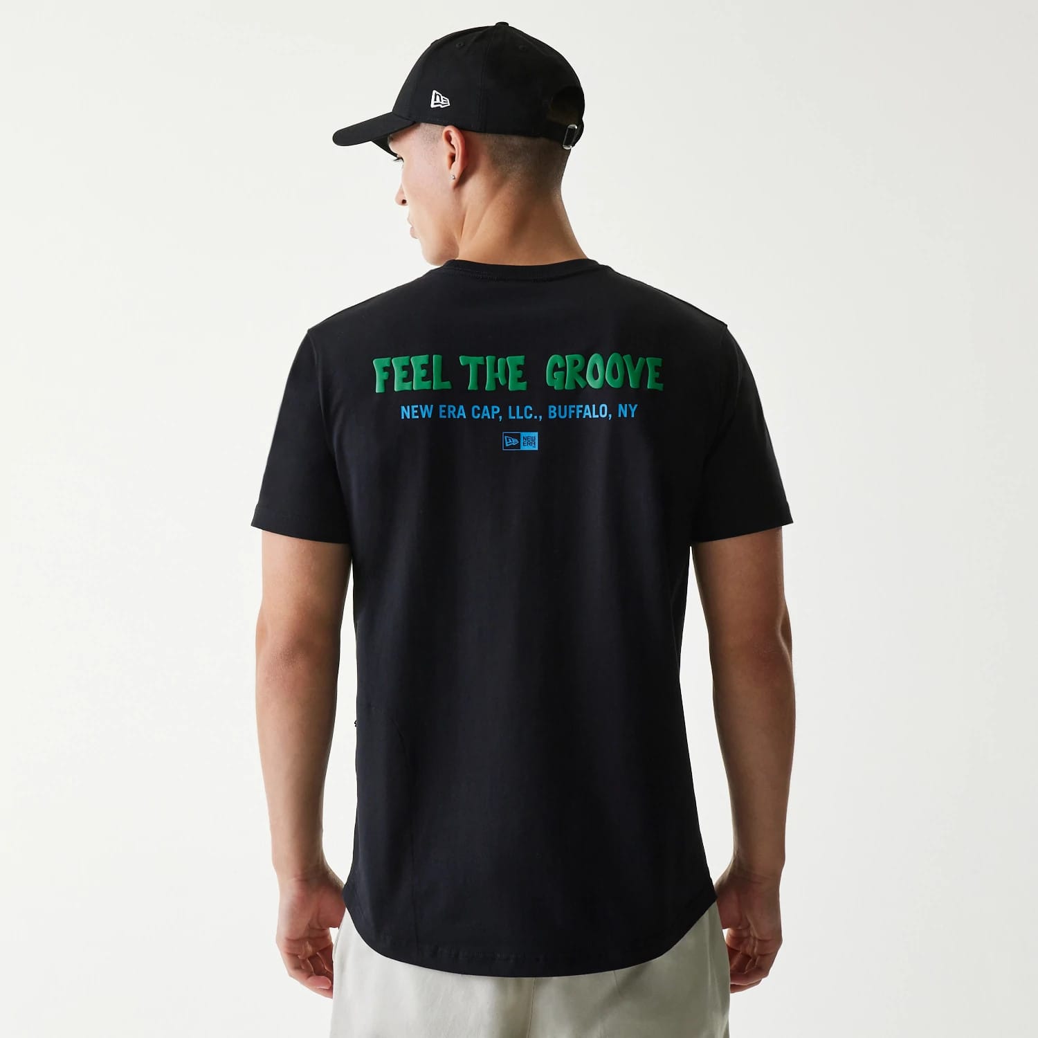 New Era Outdoor Earth Day Black Side Pocket T-Shirt, on-model view showing the back with green text "FEEL THE GROOVE" and blue New Era details below, the model wears a black cap with a debossed New Era logo on the side.