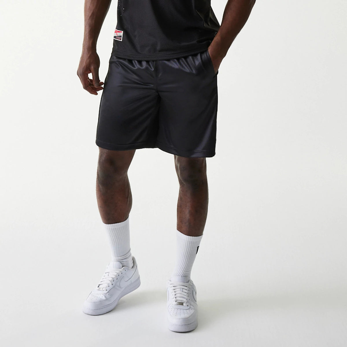 New Era Retro Sports Black Mesh Shorts, front view showing the relaxed fit silhouette, subtle piping detail along the hem, and smooth black fabric with a reflective finish, worn with white socks and trainers.