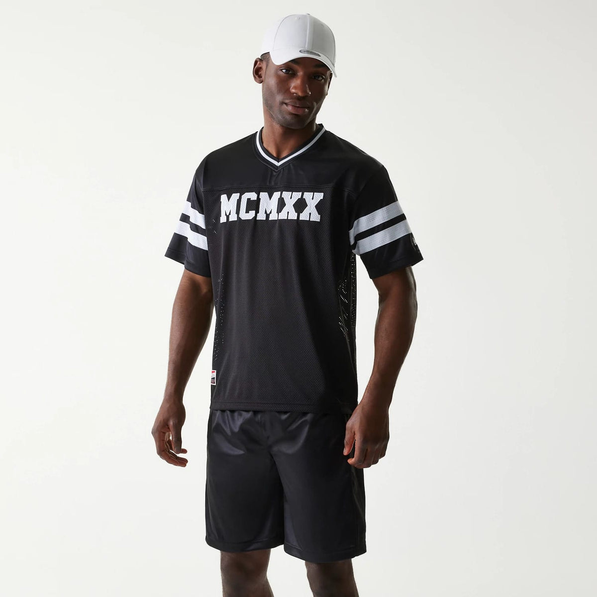 New Era Retro Sports Black Mesh Jersey, front view on a model wearing black shorts and a white cap, featuring bold white MCMXX lettering, grey and white striped sleeves and a classic V-neck design.