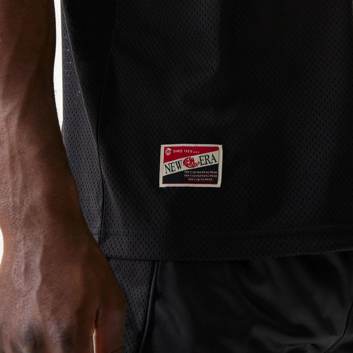 New Era Retro Sports Black Mesh Jersey, side view with a close-up of the woven vintage-style New Era label on the lower hem, the perforated mesh fabric, and a model's arm in the foreground wearing dark shorts.
