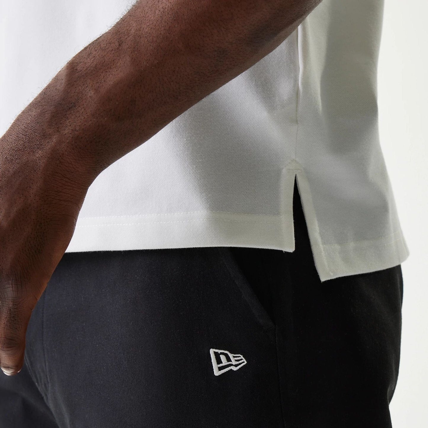 New Era Japan Collar Logo White Polo T-Shirt, side view showing the white fabric with a small side slit at the hem, accompanied by a raised New Era logo embroidered on the black trousers nearby.