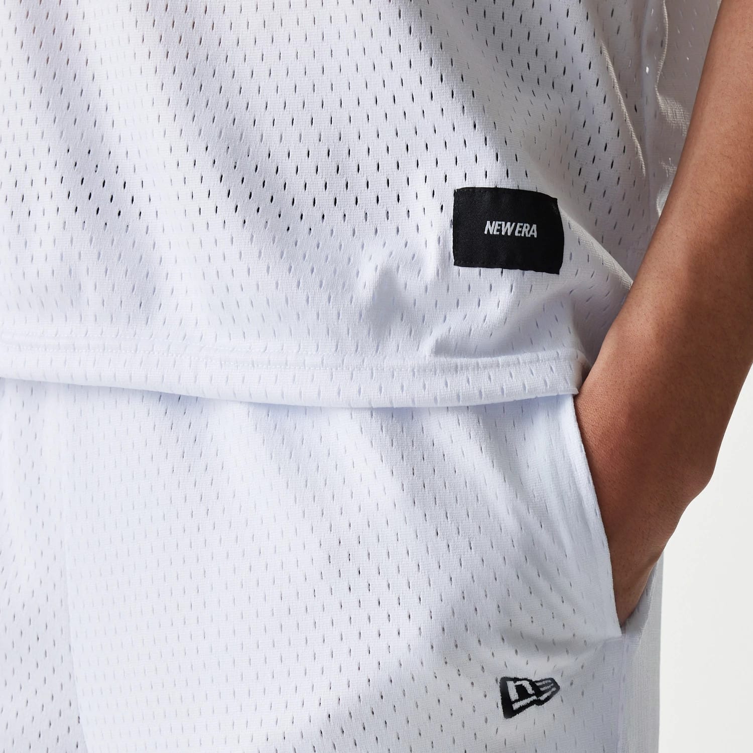 New Era Japan Mesh White Sleeveless T-Shirt, side view showing the textured mesh fabric with a black rectangular patch featuring the embossed New Era logo near the hem and a small raised logo on the matching white shorts.