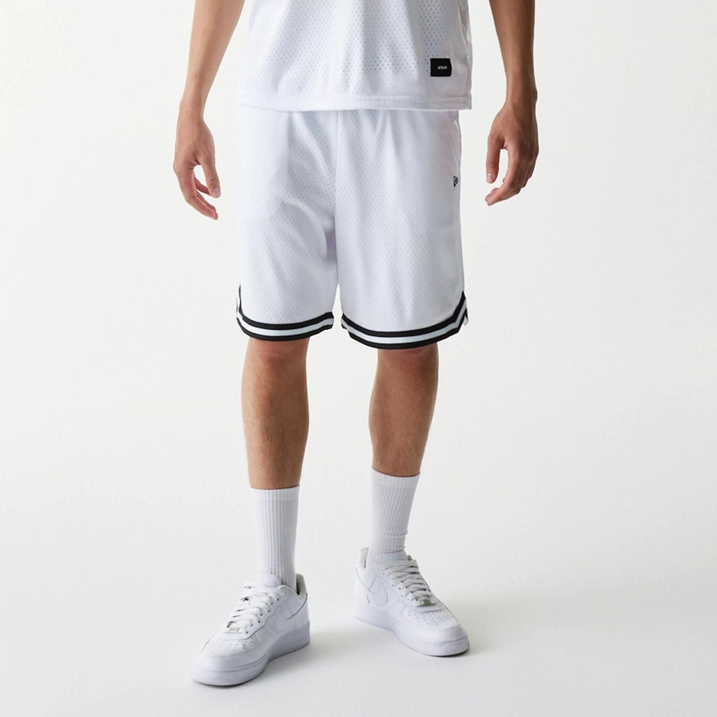 New Era Japan Mesh White Shorts, on-model view showing the white mesh fabric with black double stripes at the hem, a small debossed New Era logo on the left leg, and a relaxed fit extending just above the knee.