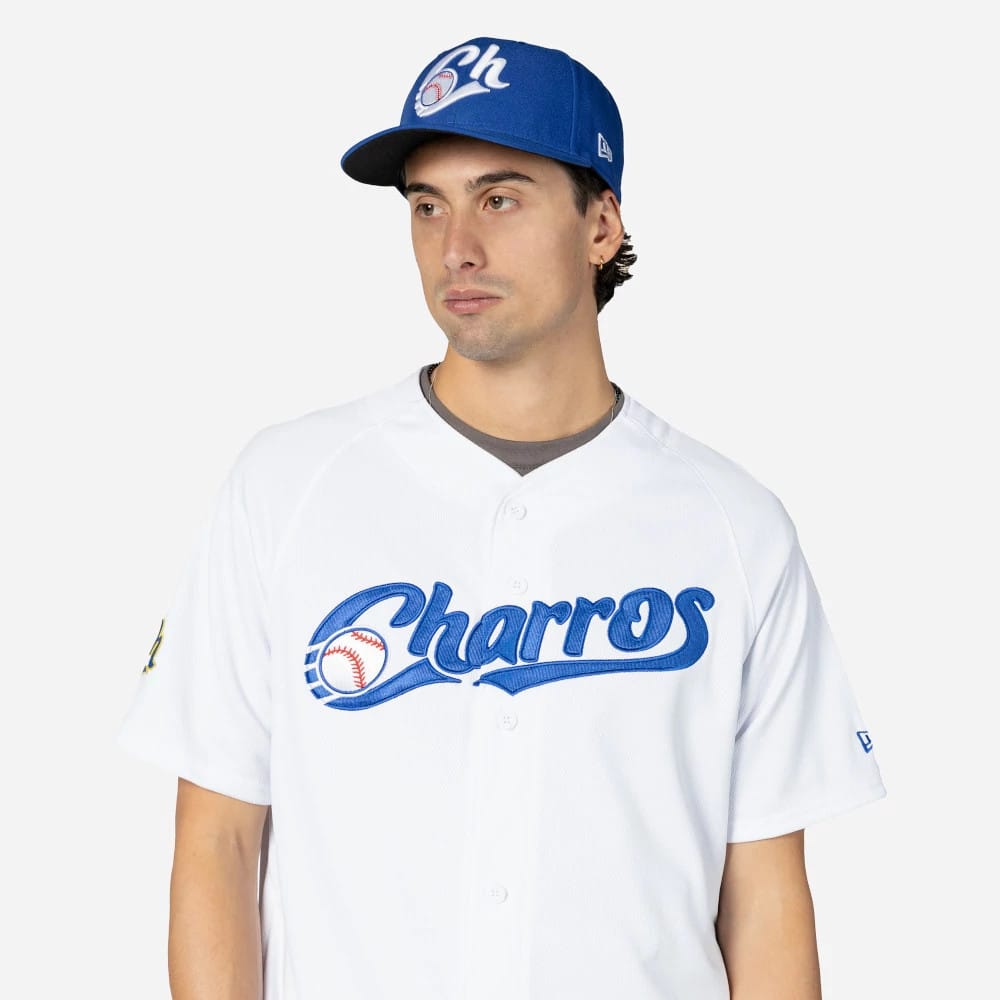 Charros De Jalisco Mexico LMB On Field White On-Field Jersey, on-model featuring large blue Charros script across the chest and a matching blue cap with the embroidered team logo, worn casually with grey undershirt visible.
