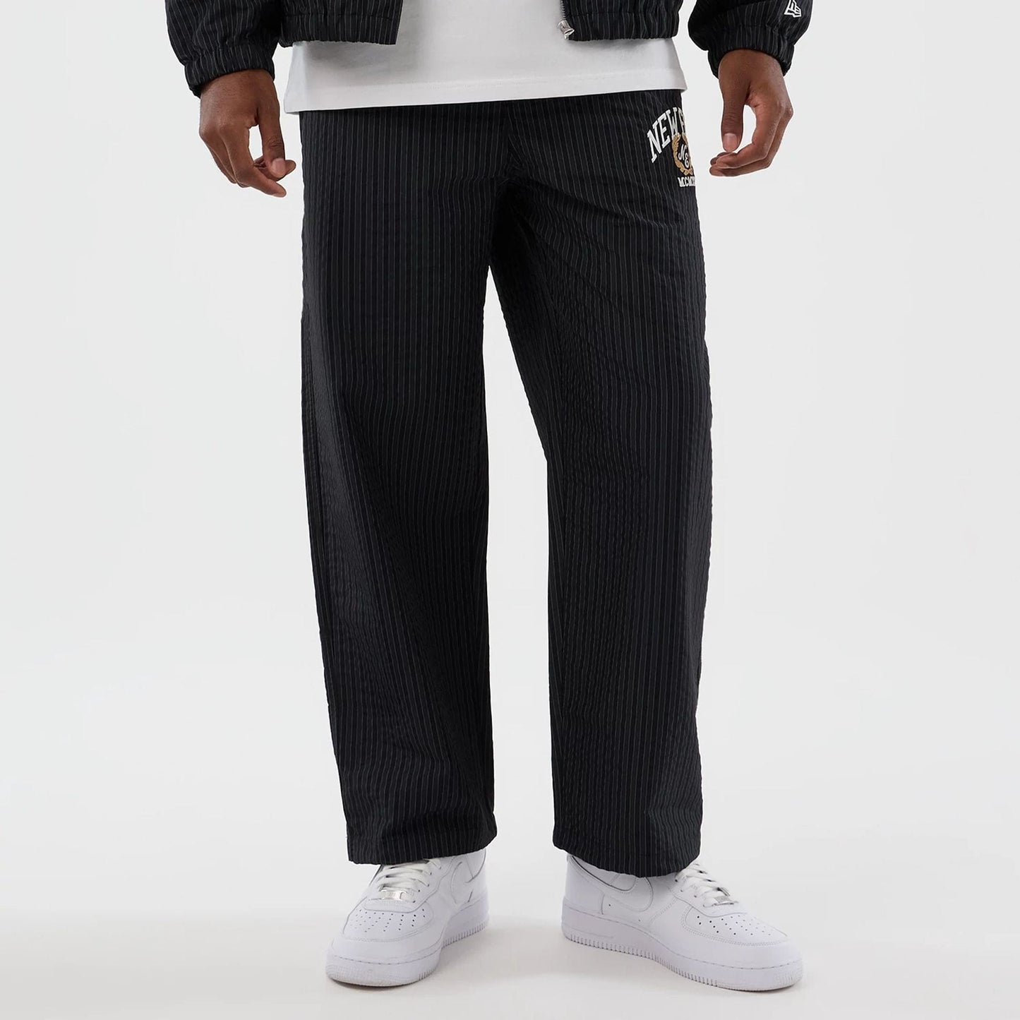 New Era Street Class Navy Straight Leg Joggers, front view on a model showing a pinstripe pattern with New York logo on the upper left thigh, styled with a matching jacket and white trainers.