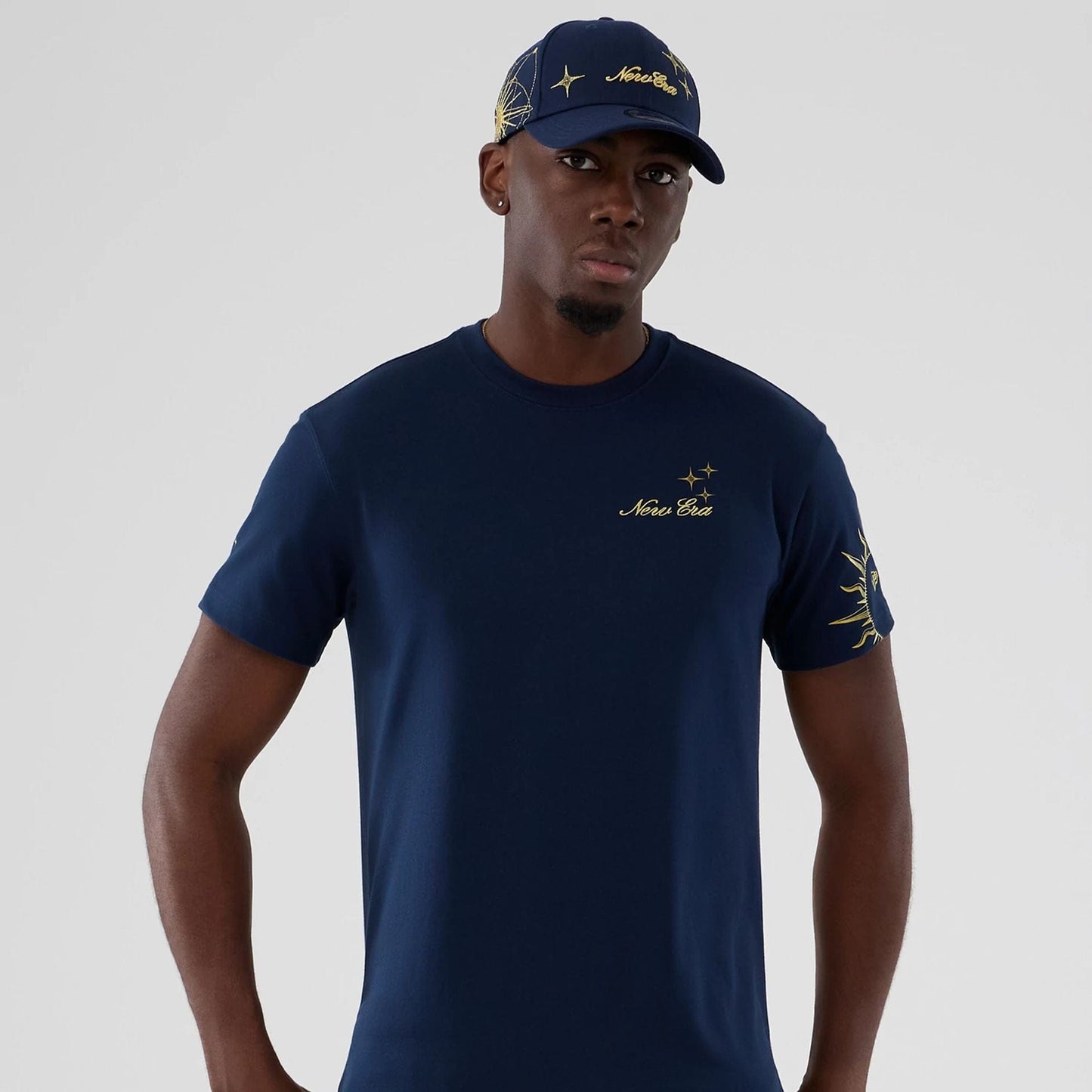 New Era Cosmic Craft Blue T-Shirt, on-model view showing the dark blue shirt with yellow embroidered New Era logo and star graphics on the chest and sleeve, paired with a matching cap featuring similar embroidery.