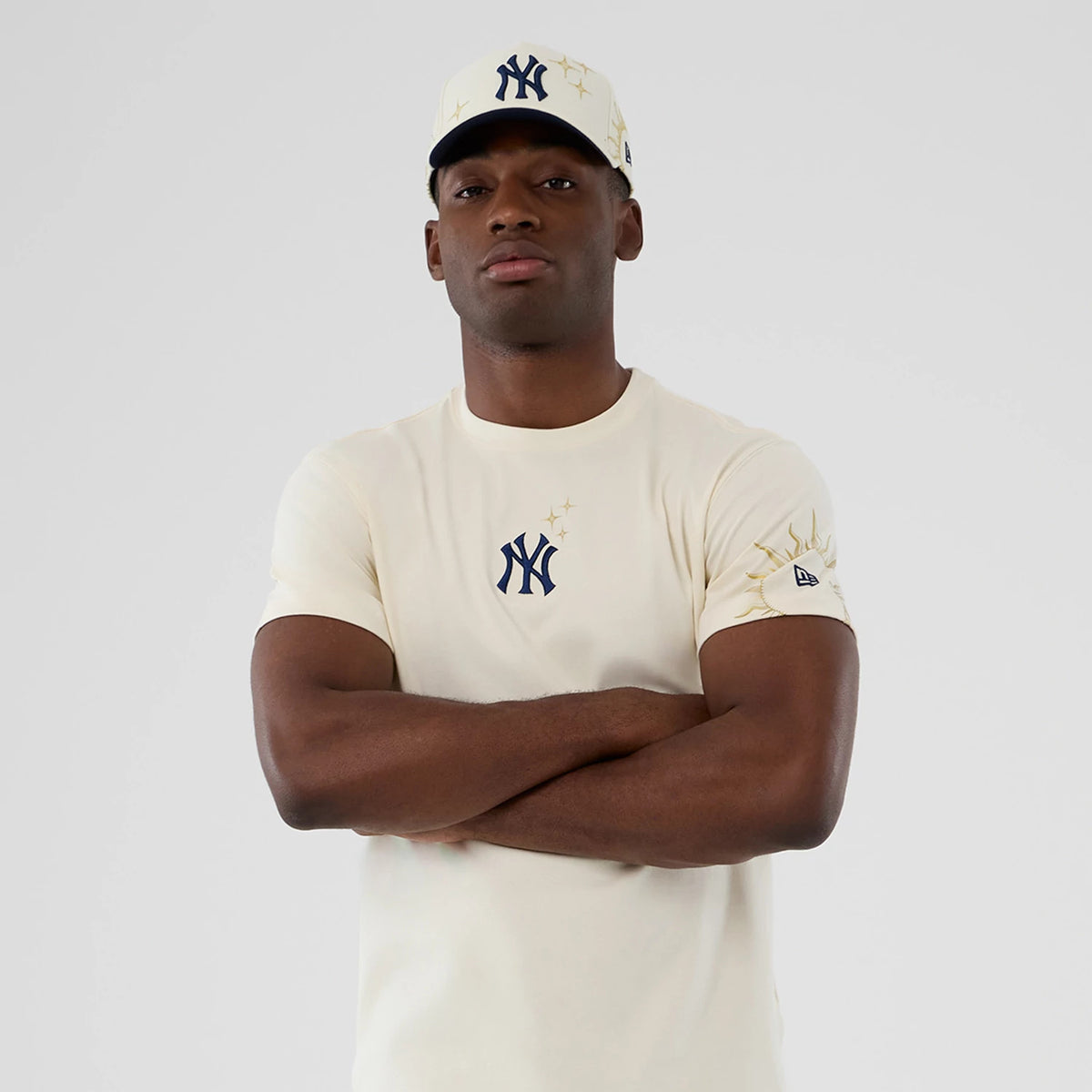 New York Yankees MLB Cosmic Craft Cream T-Shirt, on-model view showing the navy Yankees logo on the chest, with an embroidered sun design on the right sleeve and a debossed New Era logo underneath it, worn with a matching cap.