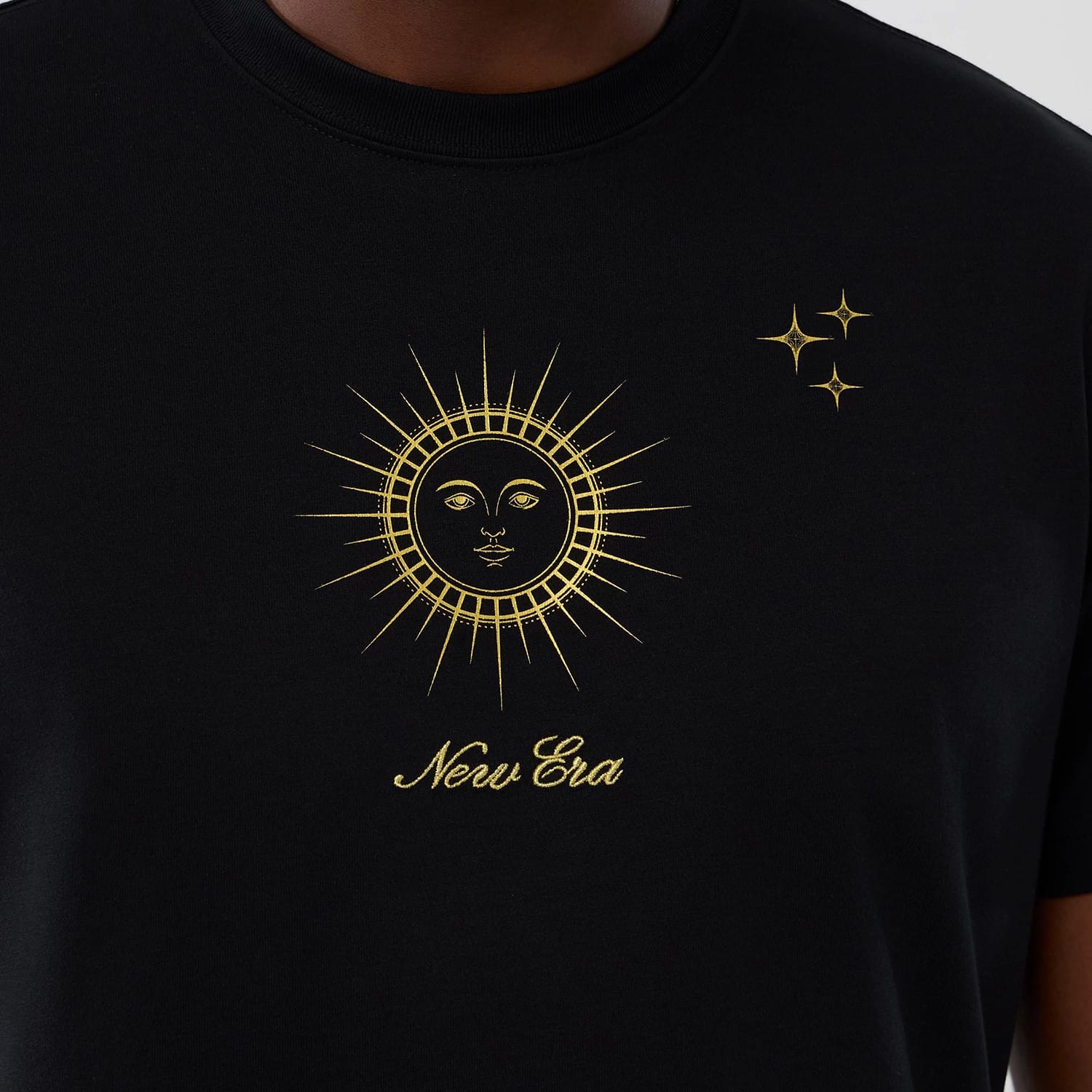 New Era Cosmic Craft Black T-Shirt, front view showing a gold sun and face design centred on the chest, a small group of gold stars to the upper right, and the New Era name embroidered in gold beneath the sun.
