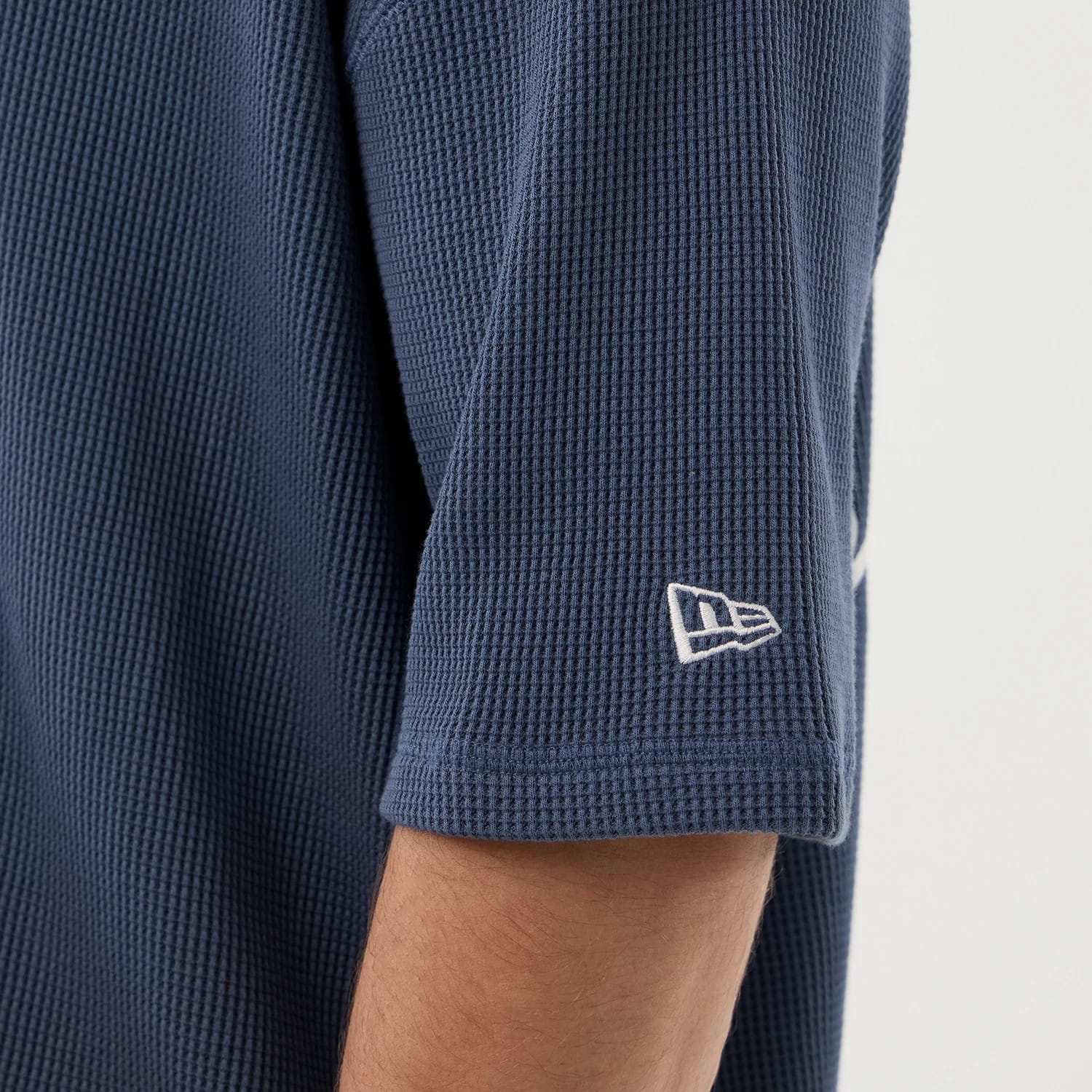 New Era Basic Blue T-Shirt, side view showing the textured blue fabric of the sleeve with a white embroidered New Era logo near the hem, displaying the clean edge and minimal design of the garment.