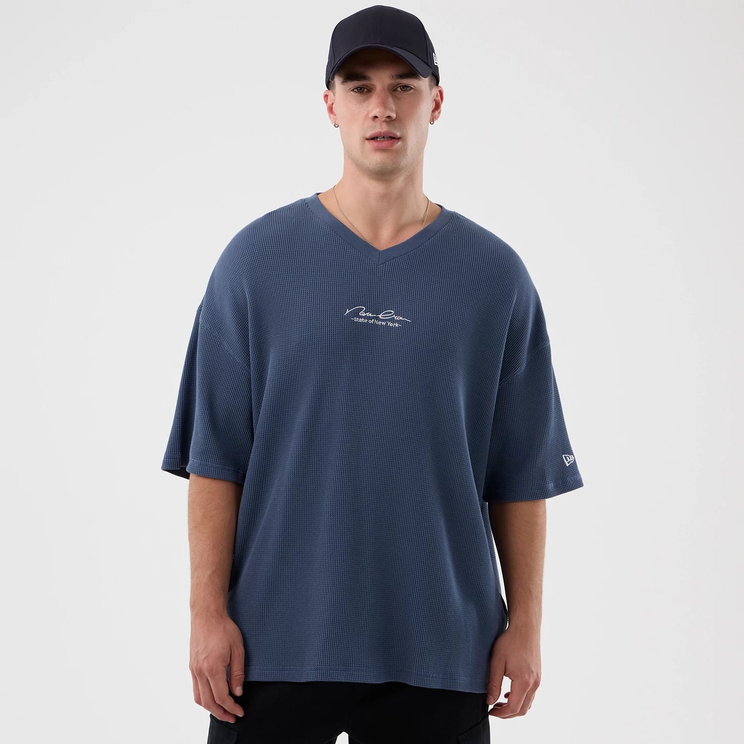 New Era Basic Blue T-Shirt, on-model view showing a loose-fitting blue V-neck T-shirt with a white embroidered signature on the chest and a small raised New Era logo on the left sleeve.