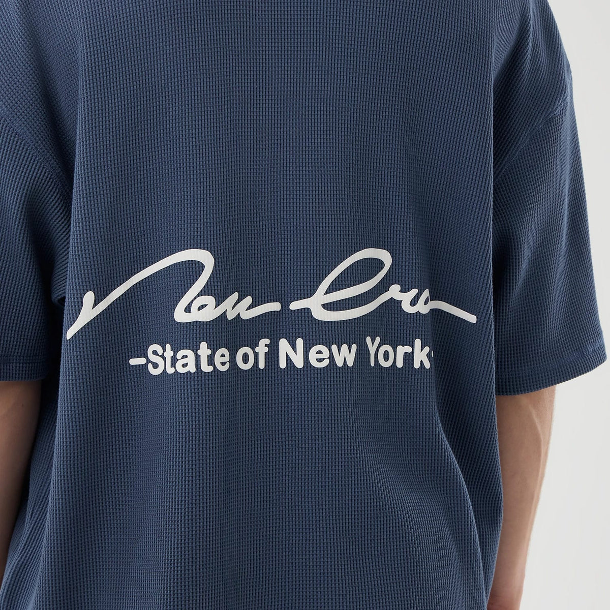 New Era Basic Blue T-Shirt, rear view showing the white printed script logo with “State of New York” text across the upper back on a textured blue fabric, short sleeves extending to the sides.