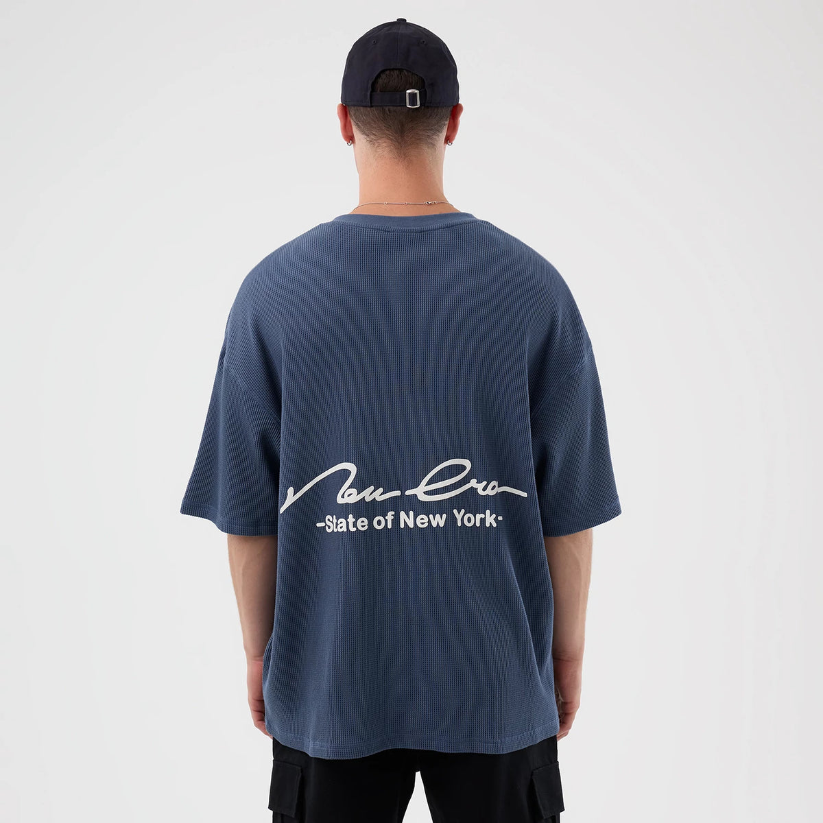 New Era Basic Blue T-Shirt, rear view showing a loose fit with a large white script New Era logo and "State of New York" printed across the back above the hem, worn by a model in a dark cap.