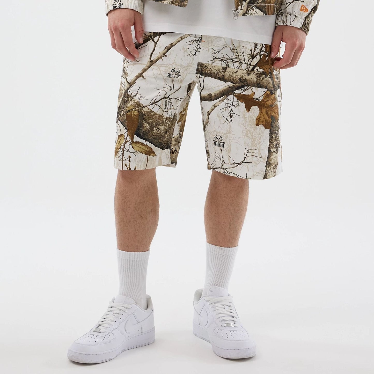 New Era Earthy Skin Real Tree Snow Oversized Shorts, front view showing the naturalistic tree and branch pattern on the white fabric, embroidered logos scattered throughout, and reaching just above the knees of the model wearing white trainers.