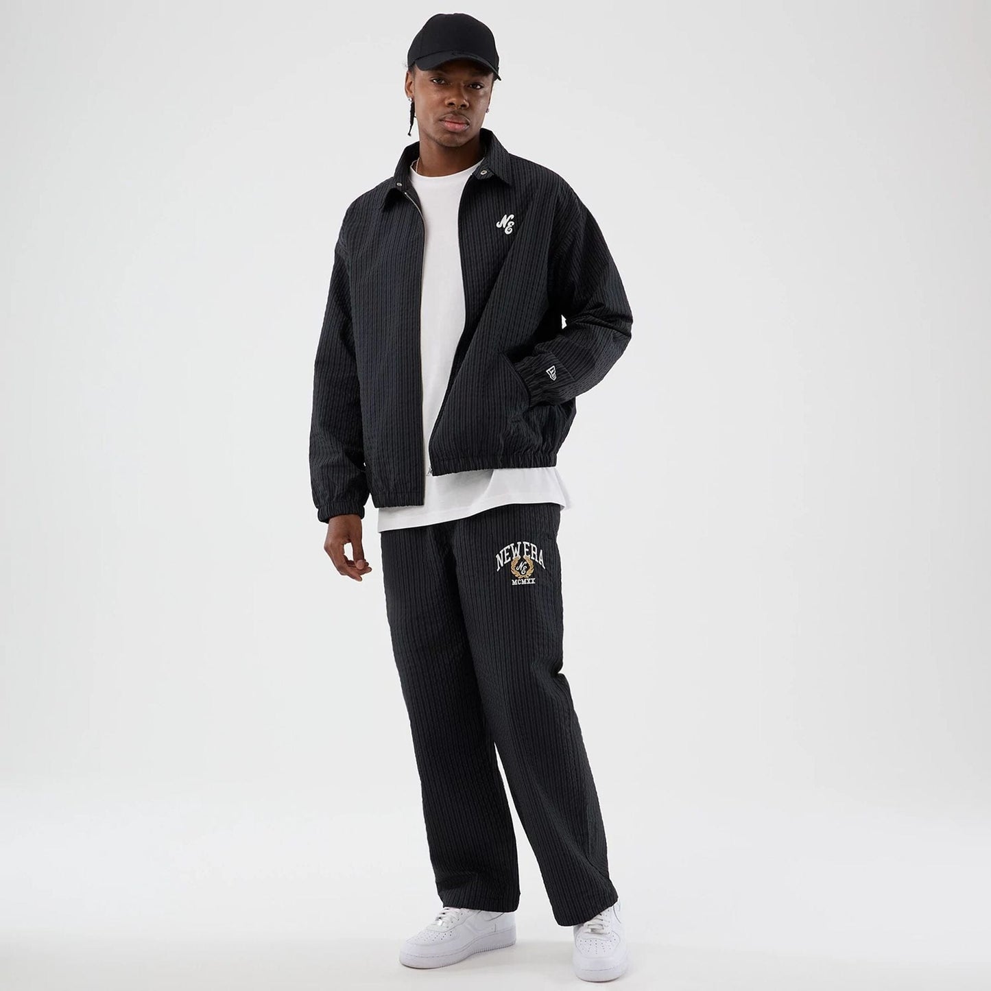 New Era Street Class Navy Jacket, on-model view showing the navy jacket with a white debossed logo on the chest, paired with matching trousers featuring a large logo on the left thigh and worn with a black cap and white trainers.