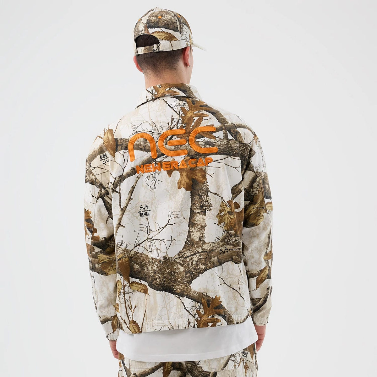 New Era Earthy Skin Real Tree Snow Jacket, rear view showing a camo pattern with brown branches and leaves, an orange raised logo mid-back, and a matching cap on-model with similar design elements.