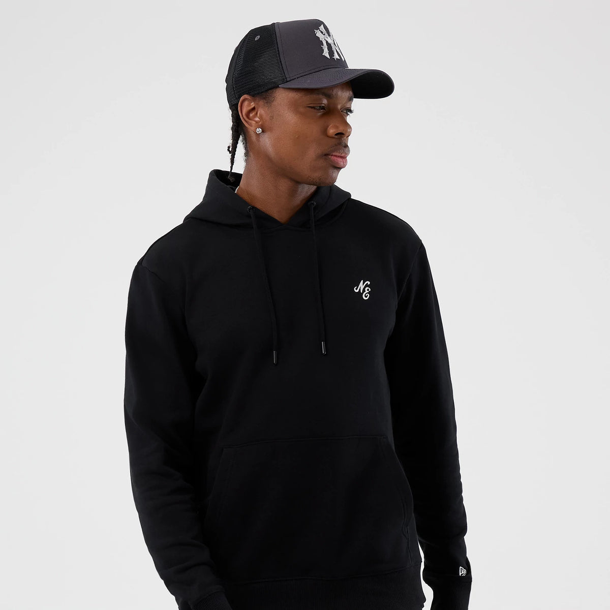 New Era Street Graphic Black Pullover Hoodie, on-model view showing the black hoodie with a white embossed "NE" logo on the front chest, an adjustable drawstring hood and a kangaroo pocket, worn with a black and grey cap featuring a raised logo.