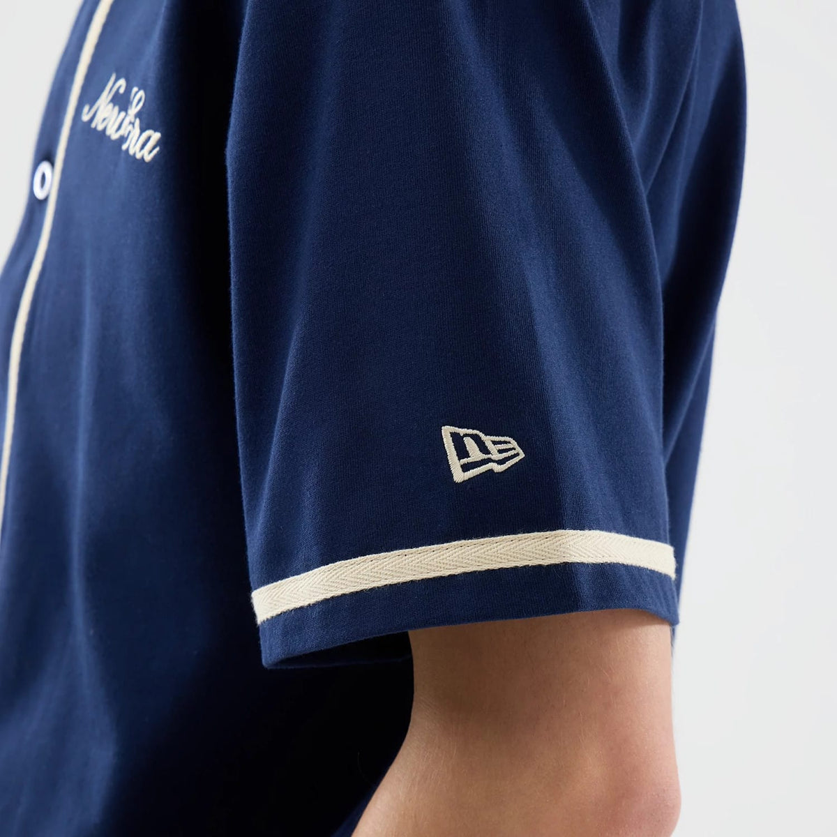 New Era Oceanside Blue Jersey, side view showing the embroidered New Era flag logo on the sleeve, with white piping along the sleeve edge and down the button placket on the deep blue fabric.