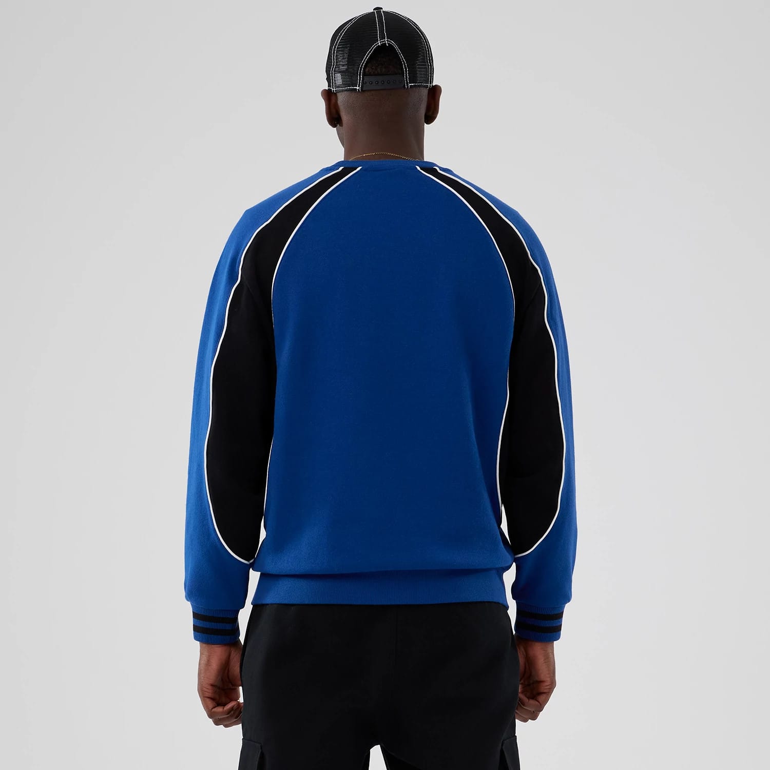 New Era Colour Block Navy Crew Neck Sweater, back view on a model showcasing blue and black panel design with white piping along the sleeves, ribbed cuffs and hem, paired with black trousers and a black cap.