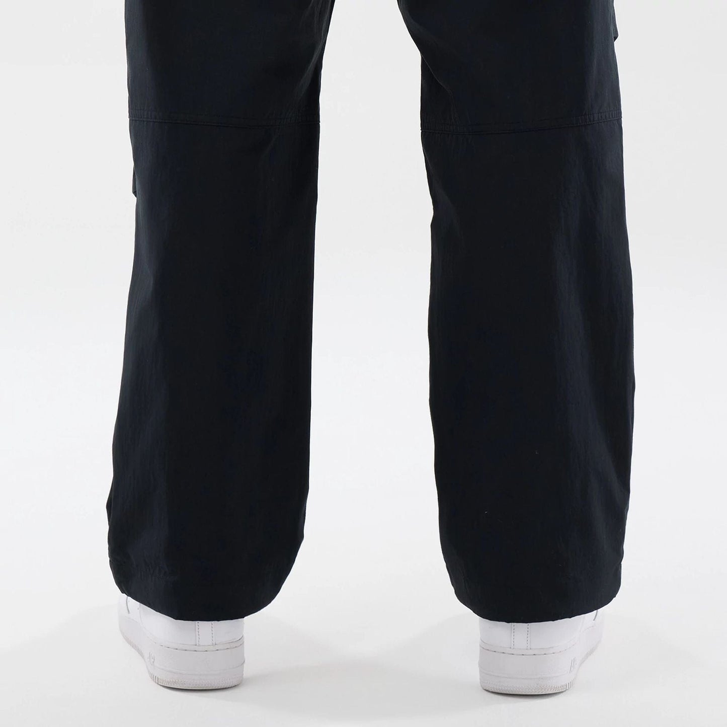 New Era Organic Black Parachute Trousers, rear view showing the loose fit draping over white sneakers, highlighting the relaxed silhouette and lightly crinkled texture of the black fabric.