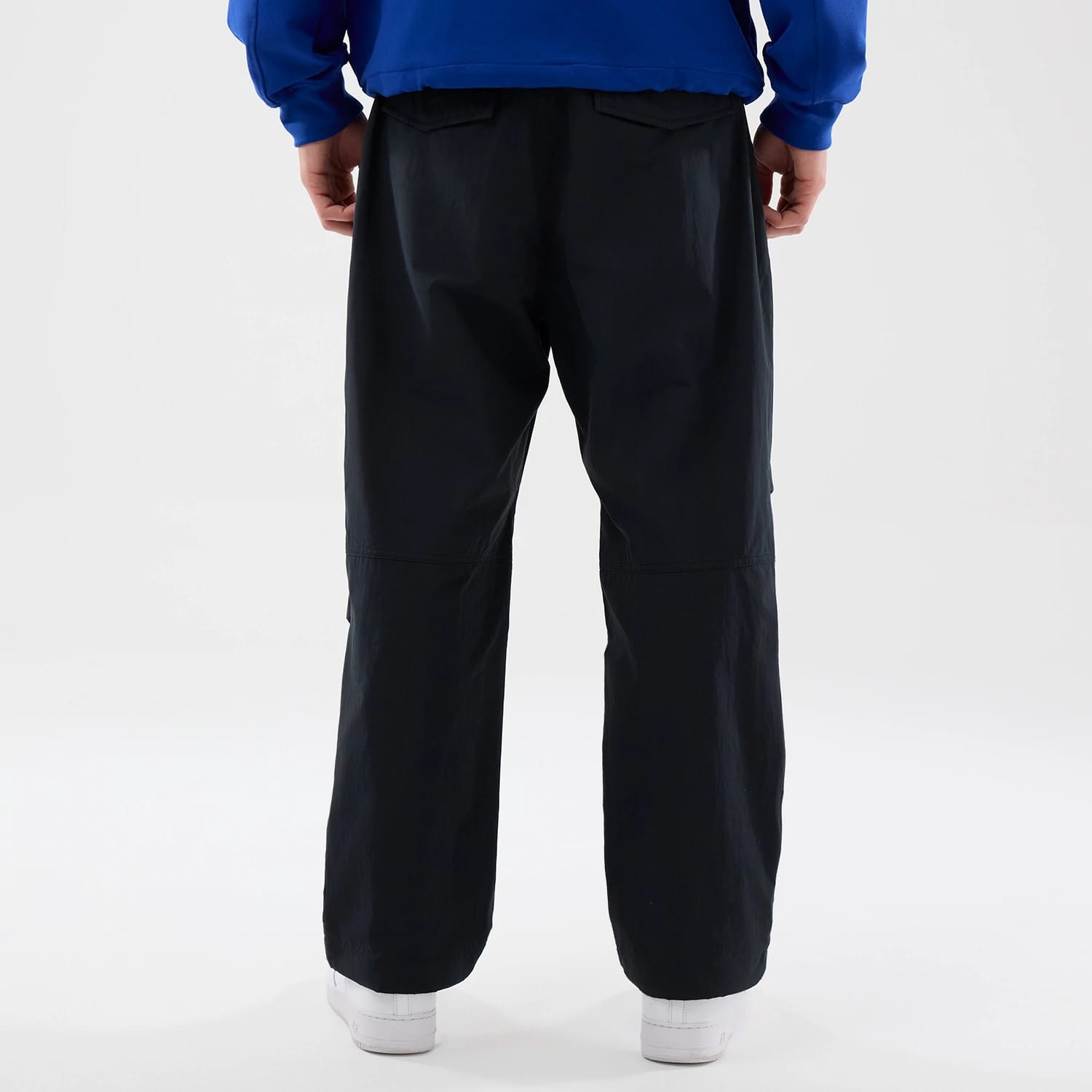 New Era Organic Black Parachute Trousers, back view on a model with a blue top and white shoes, highlighting the relaxed fit and flap pockets on the rear, showcasing the trousers' lightweight design and loose silhouette.