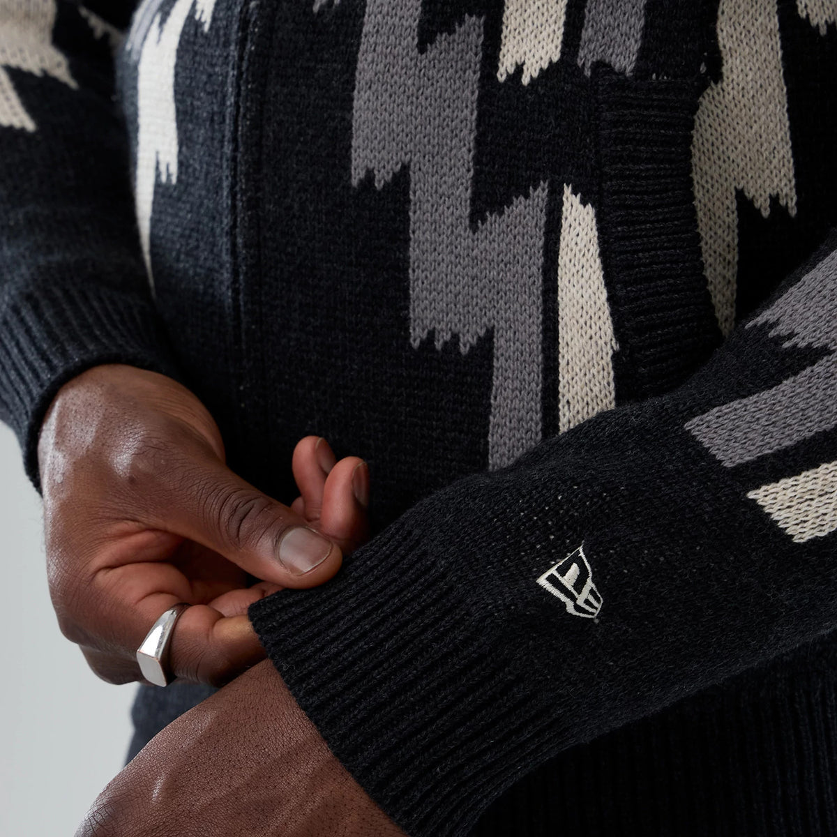 New Era Organic Cowichan Black Cardigan, close-up view showing detailed woven pattern in grey and cream shades, with a ribbed sleeve cuff embroidered with the white New Era flag logo and hands adjusting the cuff.