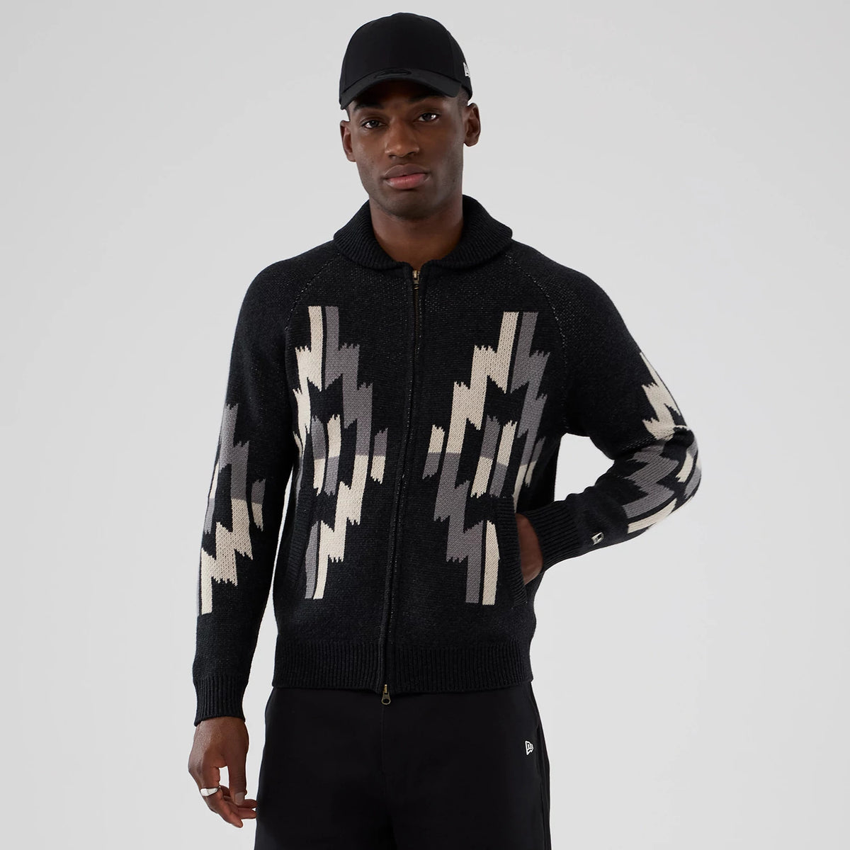 New Era Organic Cowichan Black Cardigan, front view on a model wearing a black cap, featuring a zip closure and grey geometric patterns on the chest and sleeves, with ribbed cuffs and a high collar.