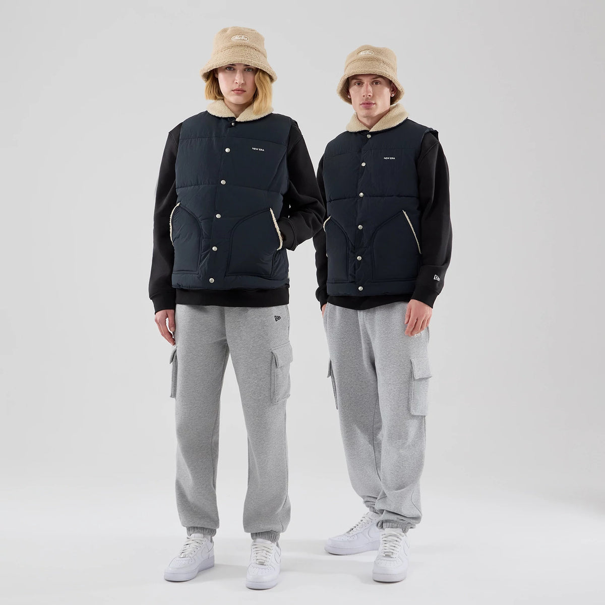New Era Organic Boa Neck Navy Gilet, front view on two models wearing beige bucket hats and grey cargo trousers, featuring a button-front closure with white buttons, cream collar, and spacious front pockets.