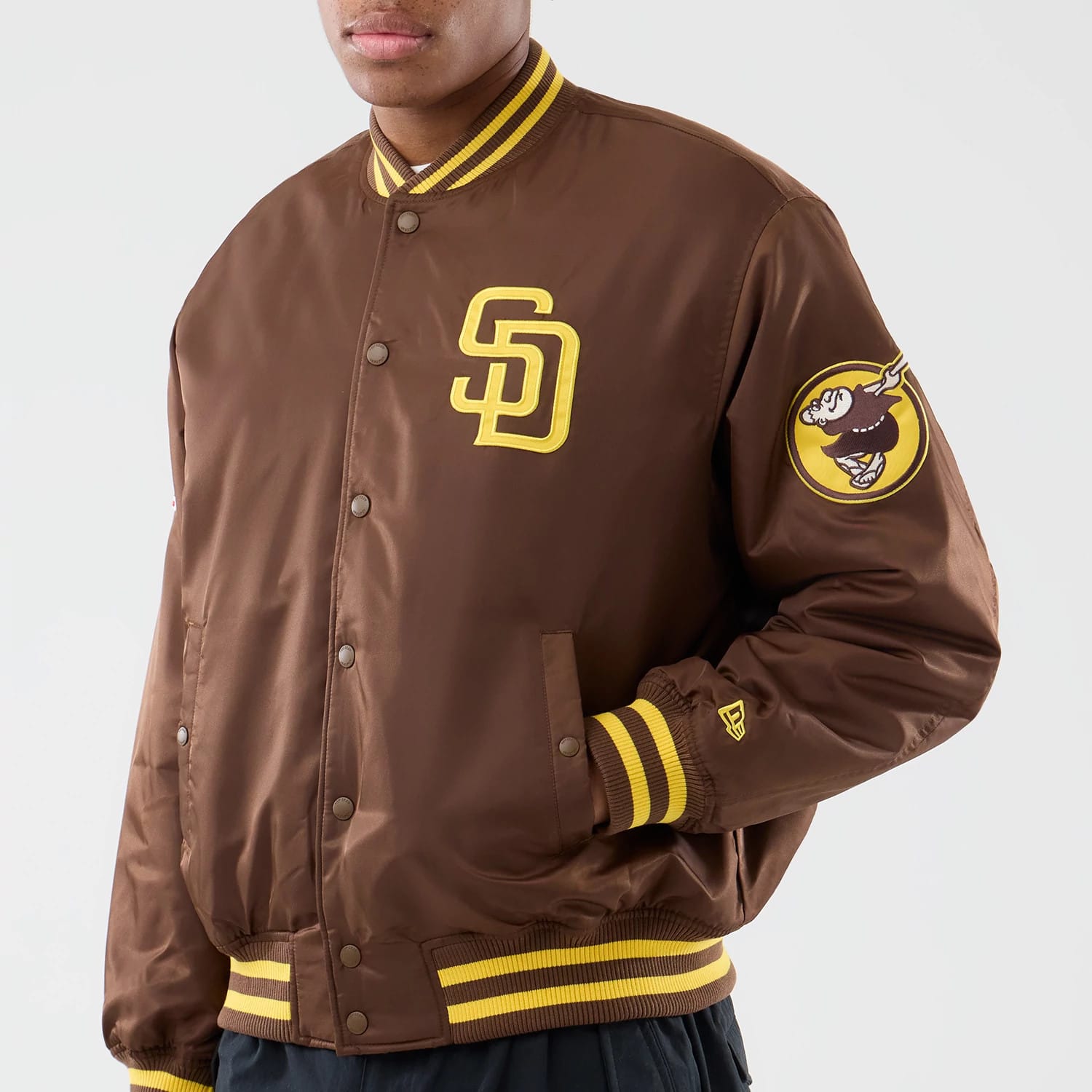 San Diego Padres MLB Authentic Dark Brown Stadium Jacket, on-model view showing a dark brown body with a raised SD logo on the chest, yellow striped cuffs and collar, a circular logo on the left sleeve, and a small raised New Era logo near the wrist.