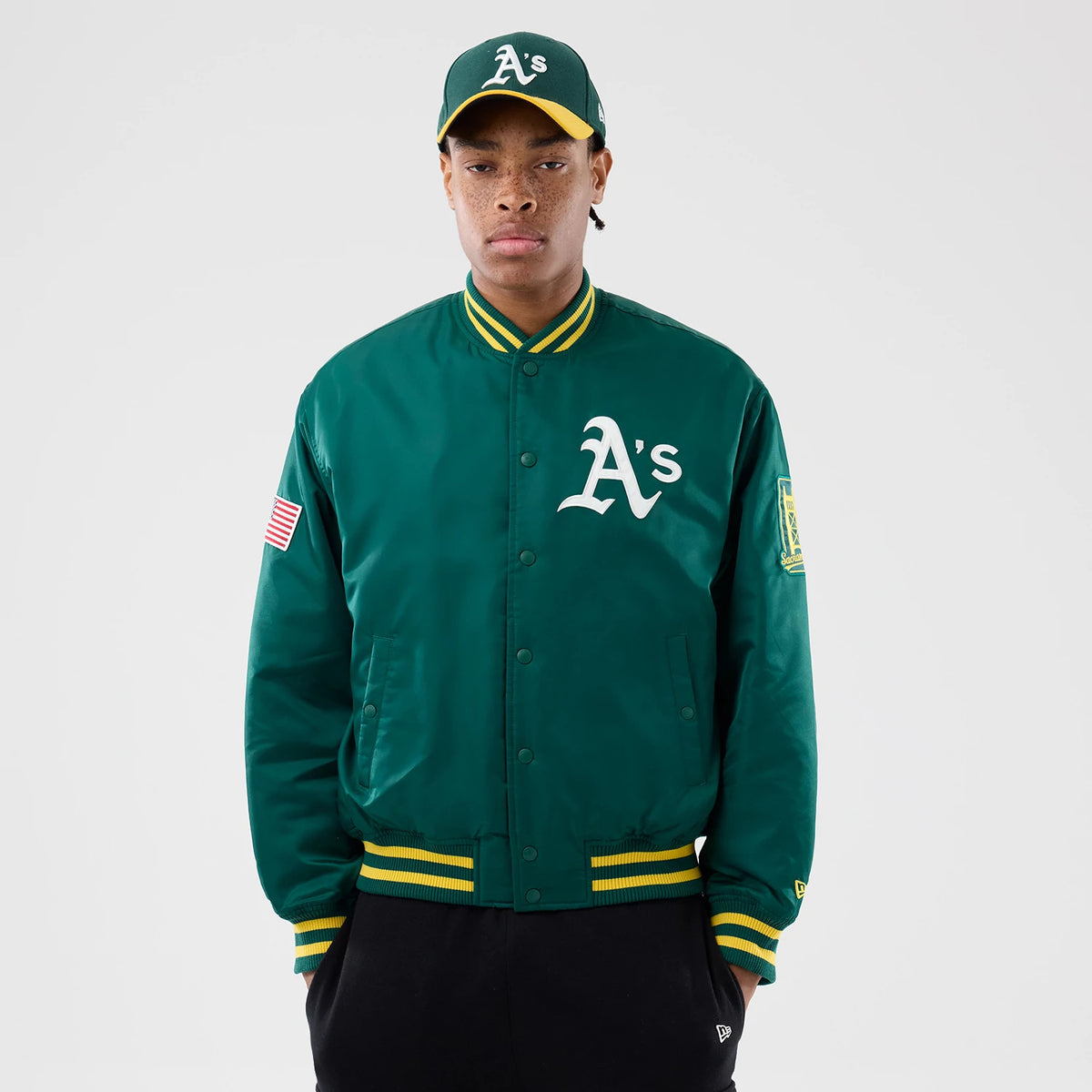 Athletics MLB Authentic Green Stadium Jacket, front view showing the green jacket with a large white A's logo on the chest, yellow and green striped collar and cuffs, a US flag patch on the right sleeve and an MLB patch on the left sleeve.