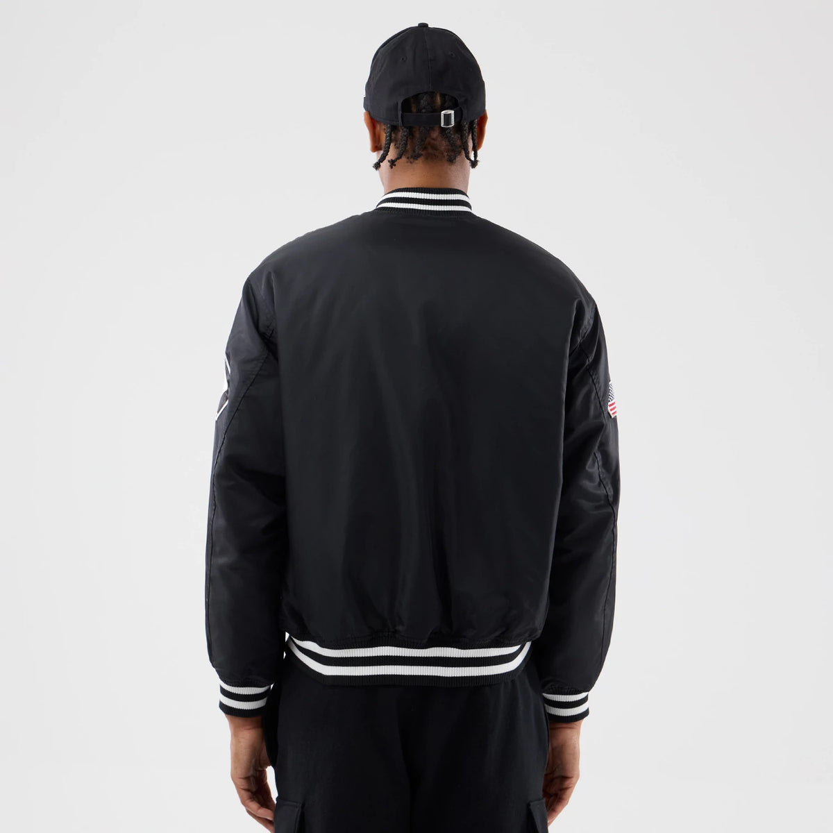 Chicago White Sox MLB Authentic Black Stadium Jacket, rear view featuring a black jacket with white striped cuffs and collar, worn by a person also wearing a black cap with a debossed logo above the back opening of the cap.