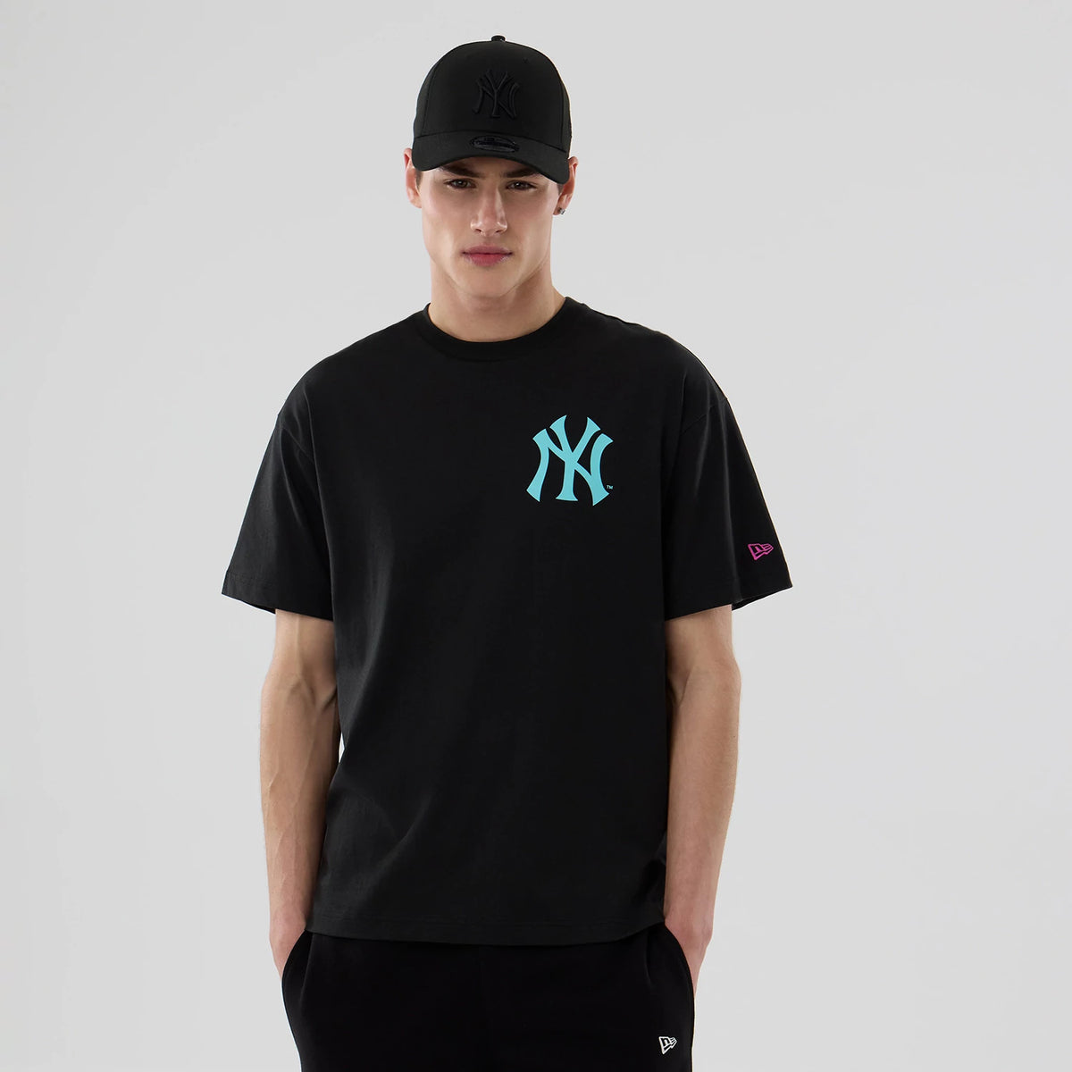 New York Yankees MLB Neon Light Black T-Shirt, on-model view showing the turquoise NY logo on the chest, a pink embroidered New Era logo on the sleeve, and a black cap with a raised NY logo worn by the model.