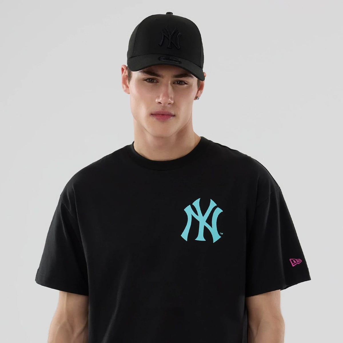 New York Yankees MLB Neon Light Black T-Shirt, on-model view showing a young man in a black T-shirt with a bright teal New York Yankees logo on the chest and a pink New Era logo on the left sleeve, paired with a black cap featuring a raised logo on the front.