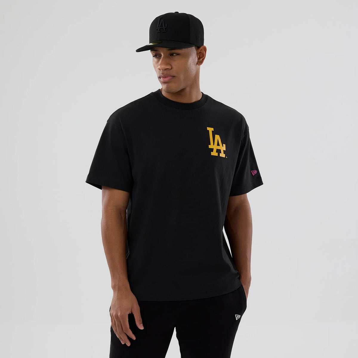 LA Dodgers MLB Neon Light Black T-Shirt, on-model view showing a large yellow LA logo on the front left of a black T-shirt, with a small magenta New Era logo on the left sleeve and an embossed black LA logo on a black cap worn by the model.