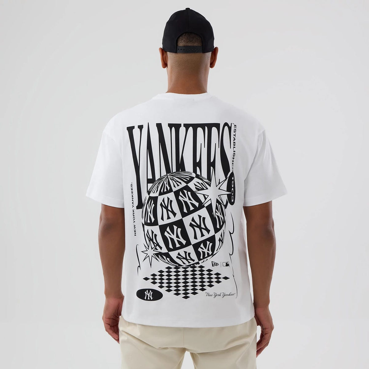 New York Yankees MLB World White T-Shirt, rear view showing a large black graphic design featuring a checkered globe with Yankees logos, bold "YANKEES" text across the back, and small logos at the bottom, worn by a person facing away.