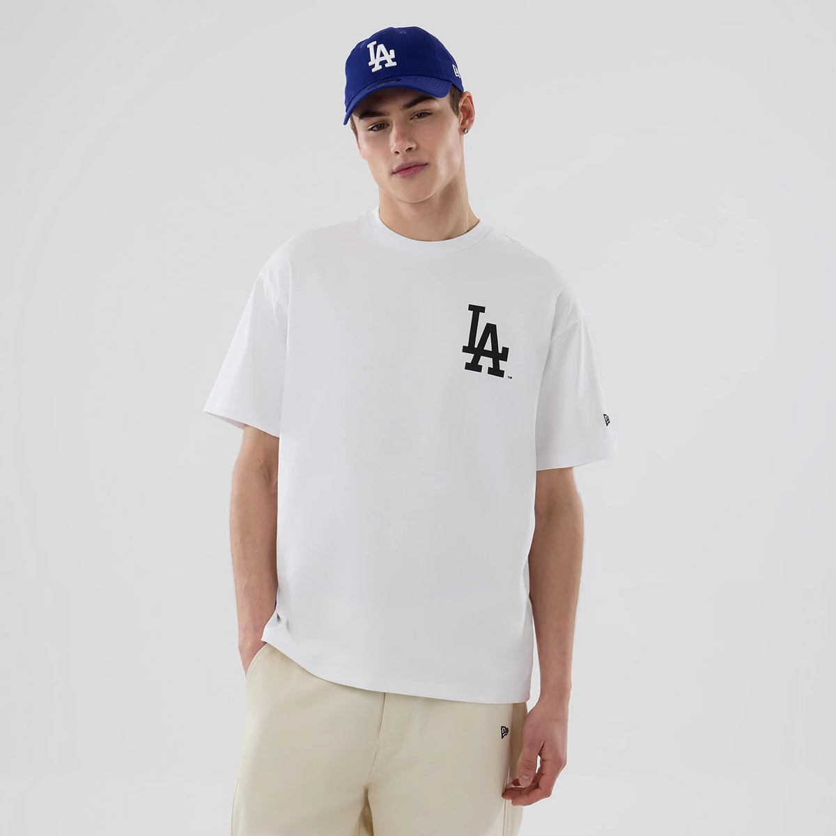LA Dodgers MLB White T-Shirt, on-model view showing the black LA logo printed on the front left side, a minimalist white fabric, and a New Era logo debossed on a blue cap worn by the model.