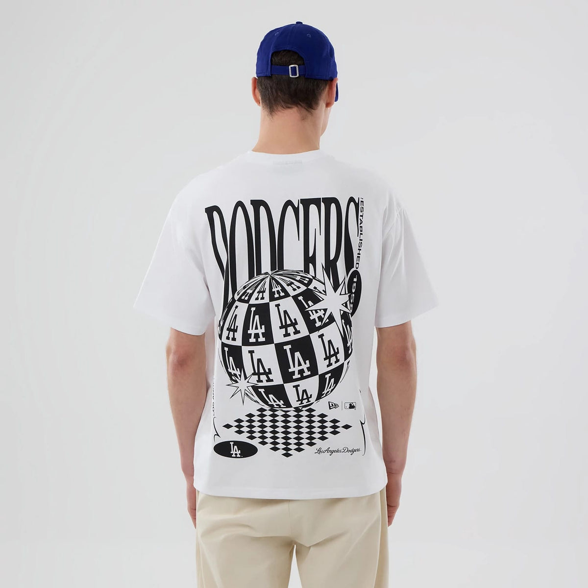 LA Dodgers MLB White T-Shirt, rear view showing large black and white graphic with repeated LA and Dodgers text, a globe design, and additional branding near the hem, worn by a person with a blue cap.