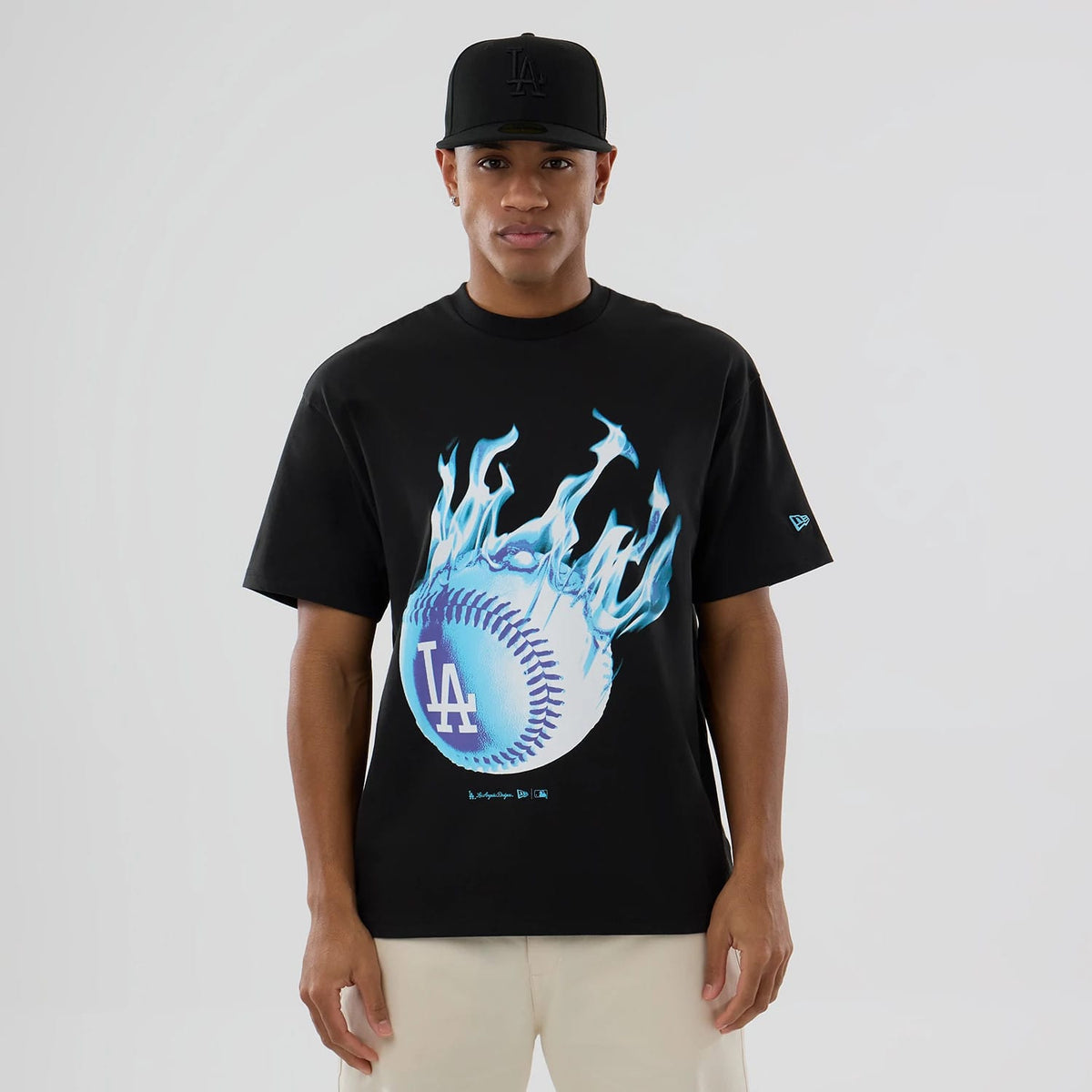 LA Dodgers MLB On Fire Black Oversized T-Shirt, front view showing a large blue flaming baseball graphic with the LA logo on the chest, a small embroidered New Era logo on the sleeve, and a person wearing a black cap with a debossed logo.