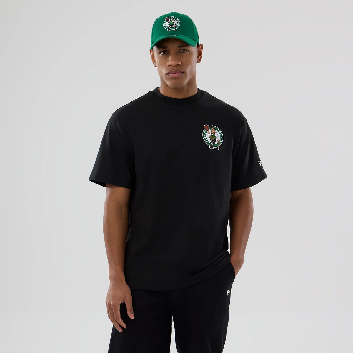 Boston Celtics NBA logo Black T-Shirt, on-model view showing the black T-shirt with a Boston Celtics logo on the left chest, a small debossed New Era logo on the left sleeve, paired with a green cap featuring the same Celtics logo.