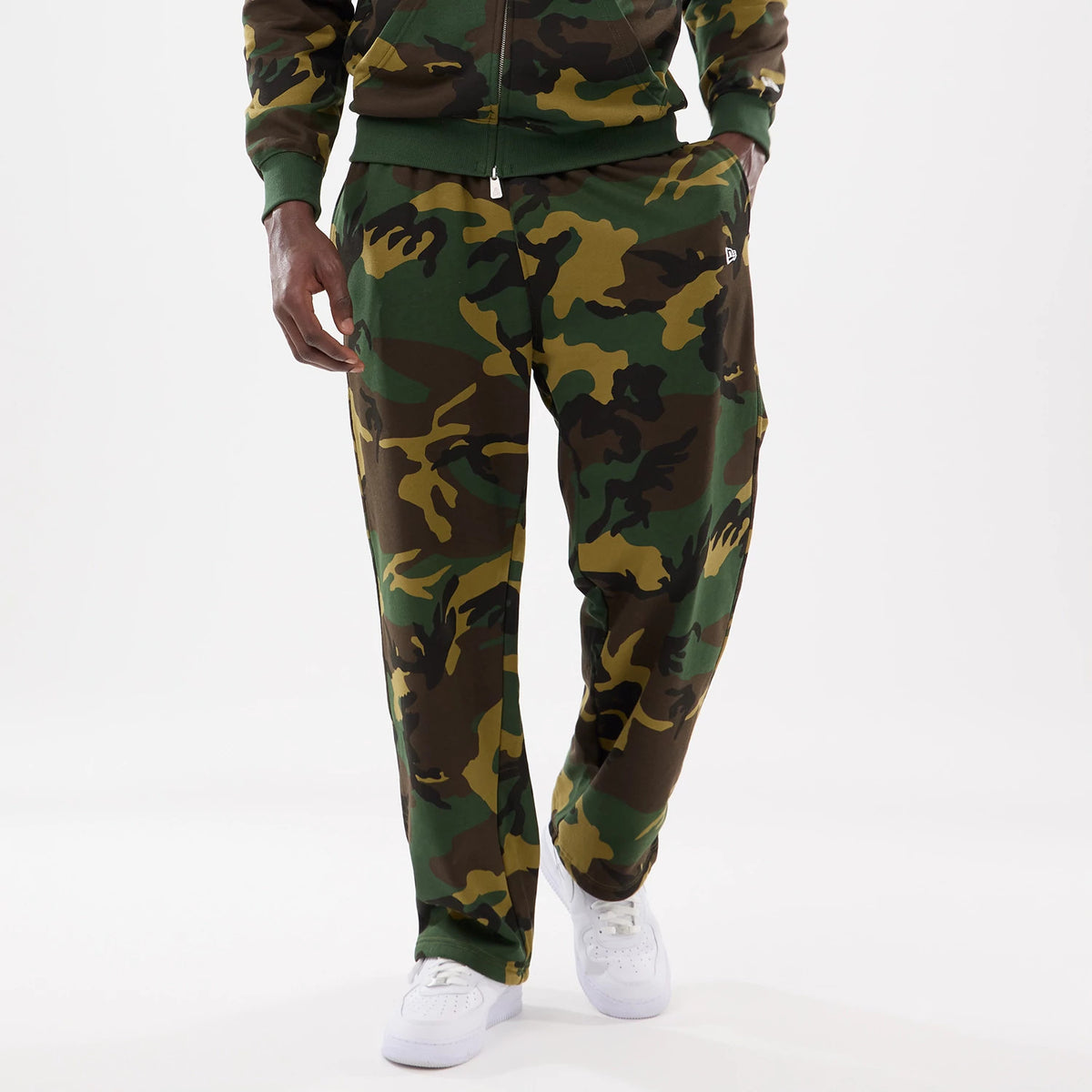New Era Woodland Camo Joggers, front view on a model showing a green, brown, and black camouflage pattern, with the New Era flag logo on the left thigh, paired with white trainers.