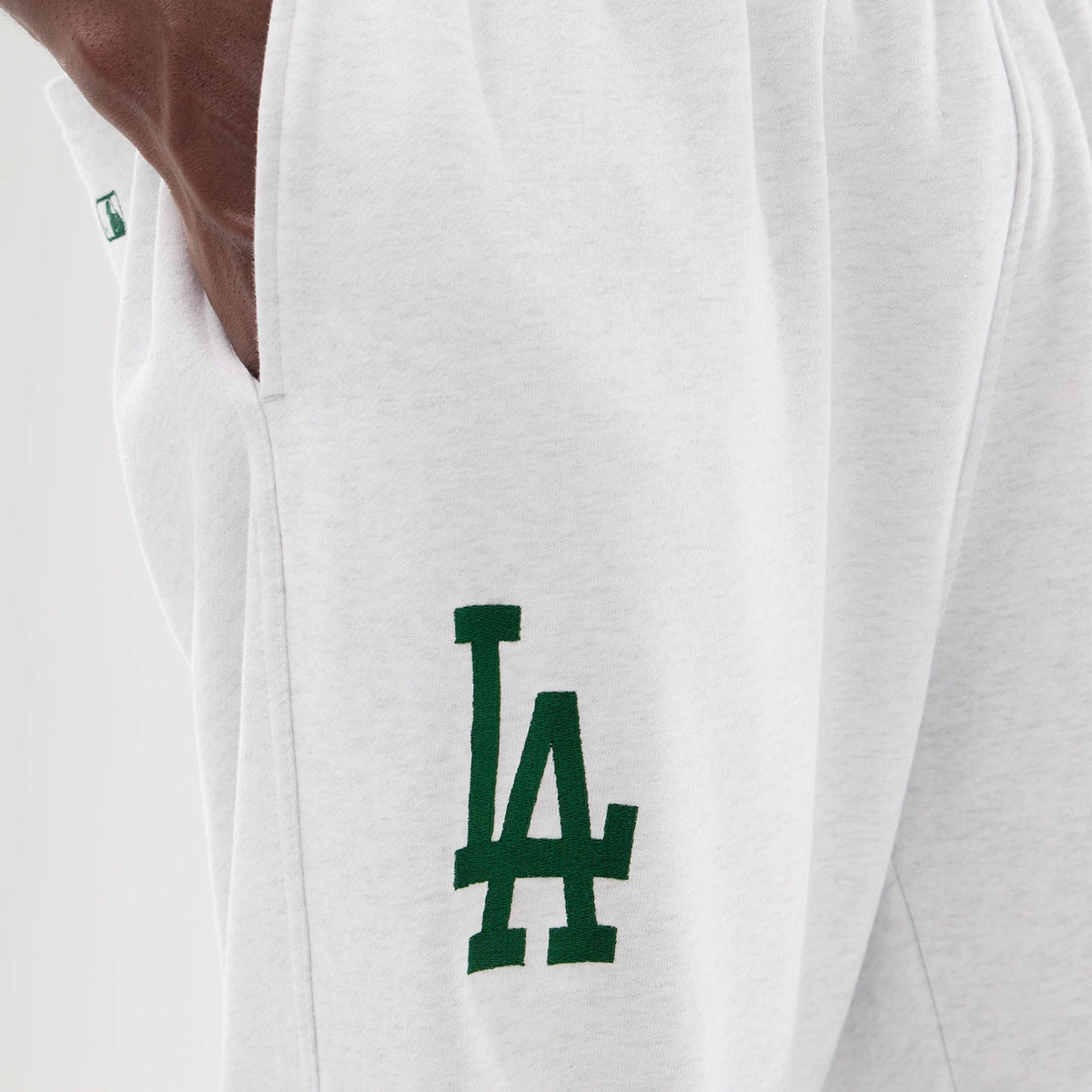 New Era LA Dodgers MLB Comfort Grey Joggers, close-up showing the dark green LA logo embroidered on the left thigh, with a small green tag on the side seam and a soft grey fabric texture.