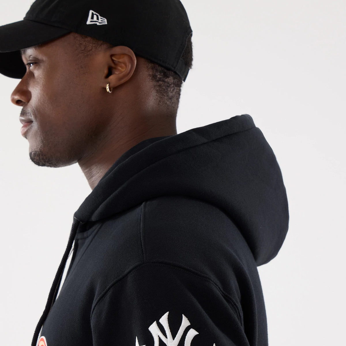New York Yankees MLB Word Art Black 2 Way Full Zip Hoodie, side view showing the black hoodie worn by a person, with a white raised New Era logo on a black cap, and white Yankees lettering down the sleeve.