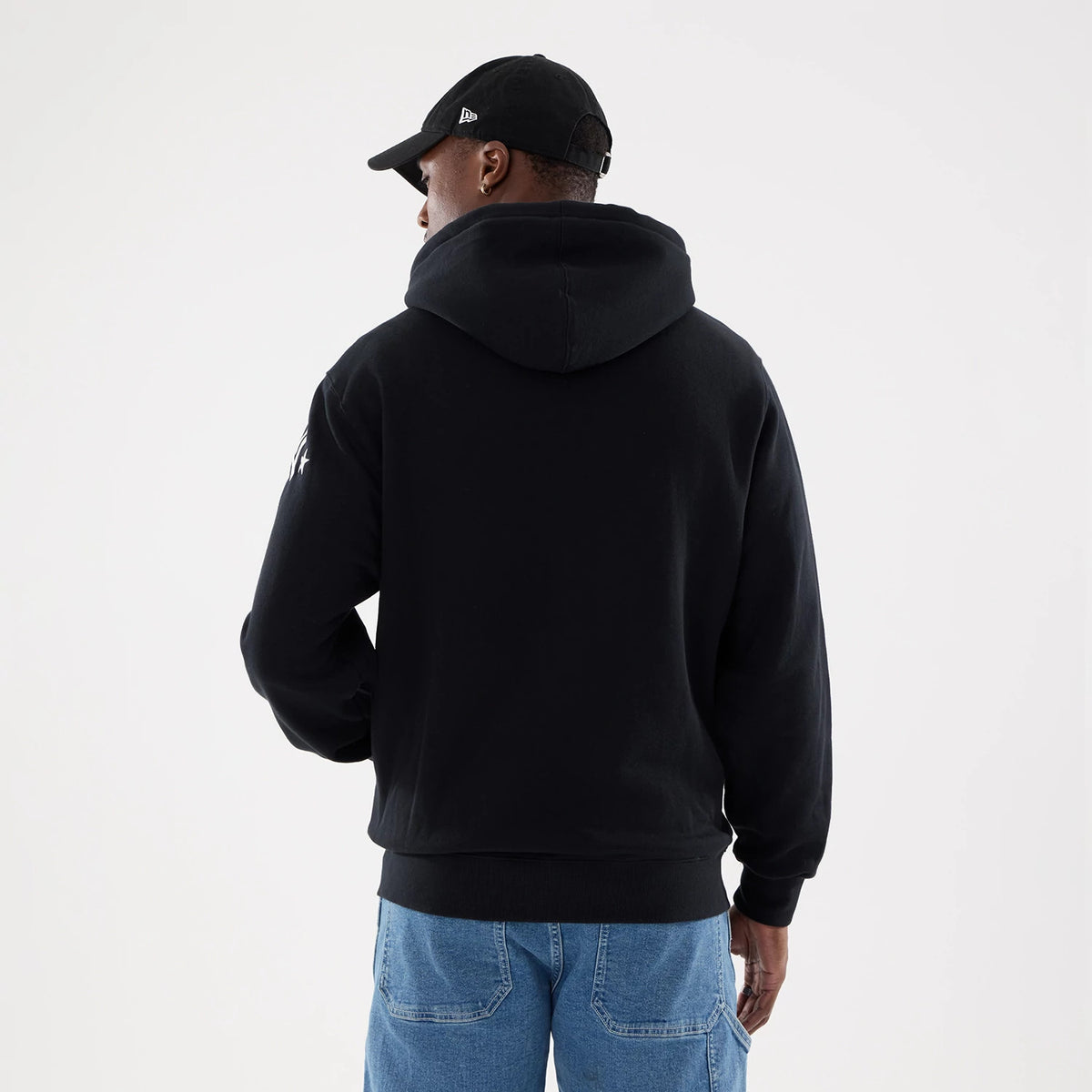 New York Yankees MLB Word Art Black 2 Way Full Zip Hoodie, rear view showing a plain black hood and a white embroidered logo on the upper right sleeve, worn by a model in a black cap with a raised logo and light blue jeans.