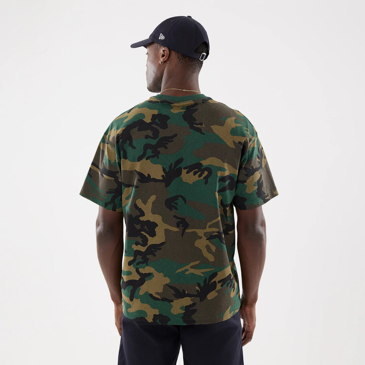 New York Yankees MLB Camo Woodland T-Shirt, rear view showing the green and brown camouflage pattern covering the entire back, short sleeves, and a crew neckline, worn by a person also wearing a black cap with a raised logo on the side.
