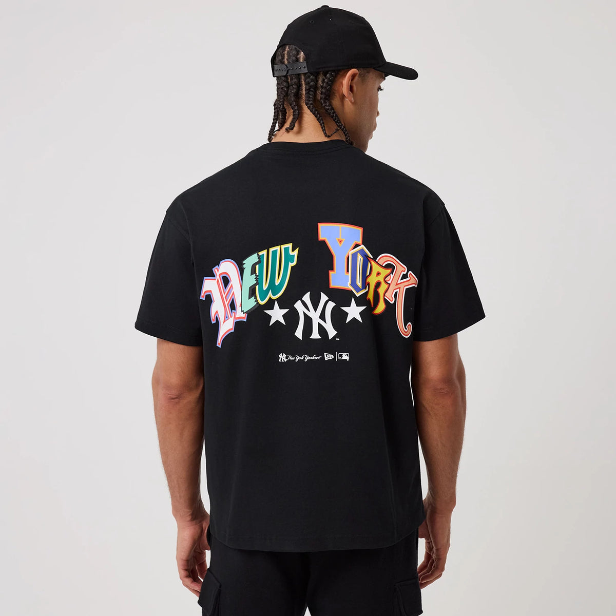 New York Yankees MLB Word Art Black T-Shirt, rear view showing multicoloured "New York" lettering with a white Yankees logo and stars on the black fabric, worn by a model in a black cap.