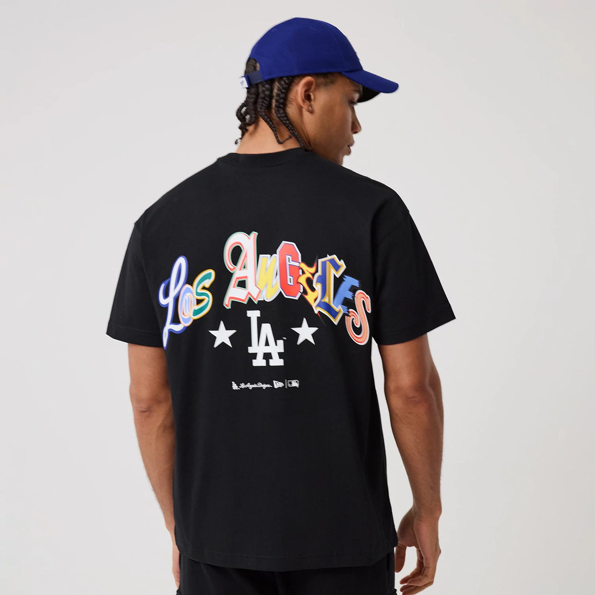 LA Dodgers MLB Word Art Black T-Shirt, rear view featuring colourful Los Angeles text across the upper back, a bold white LA logo below flanked by stars, and worn by a person in a blue cap.