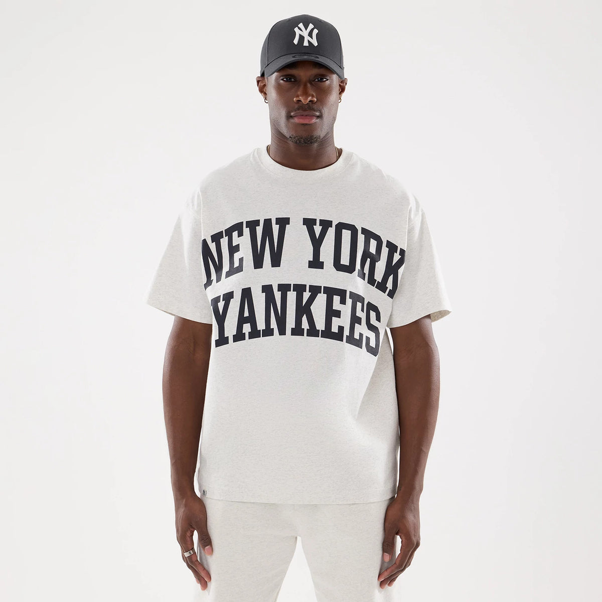 New York Yankees MLB Comfort Grey T-Shirt, front view showing the grey fabric with large black New York Yankees lettering across the chest, worn by a model, paired with a black cap featuring a raised white logo.